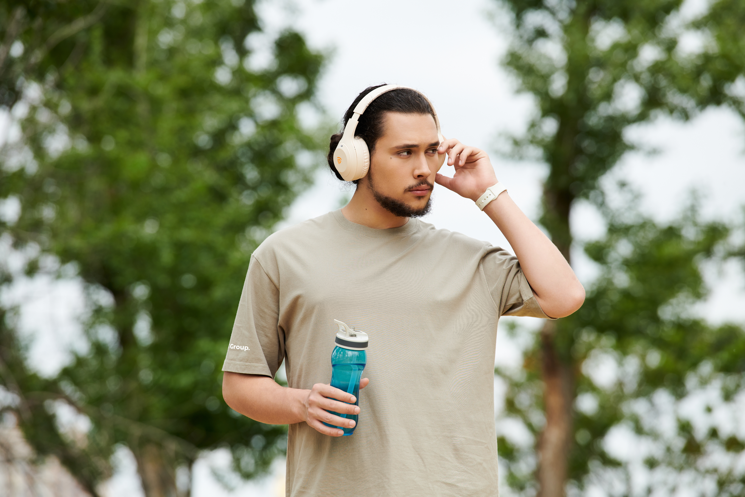A young man outdoors wearing headphones, holding a water bottle, and adjusting one earbud, surrounded by trees.