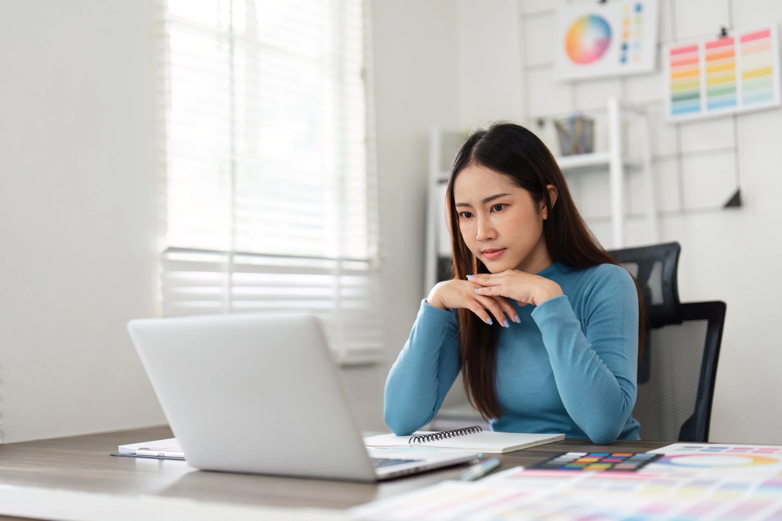 Woman in a blue shirt working at a desk with color swatches and a laptop, looking focused.
