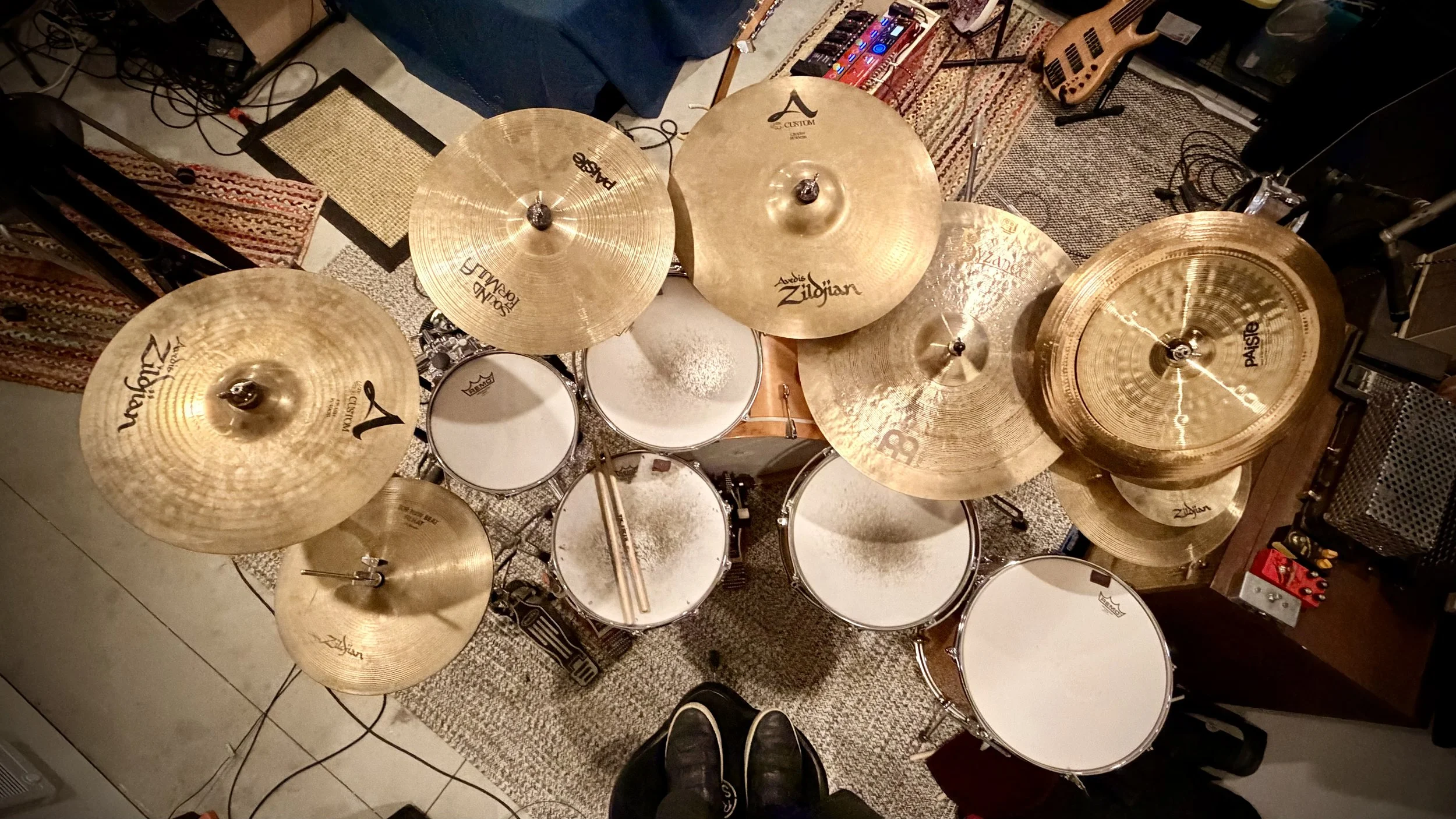 Overhead view of a drum kit with multiple cymbals, drums, and drumsticks placed across two of the drums. Surrounding the setup are various pieces of musical equipment and furniture, including rugs and an electric guitar in the background.