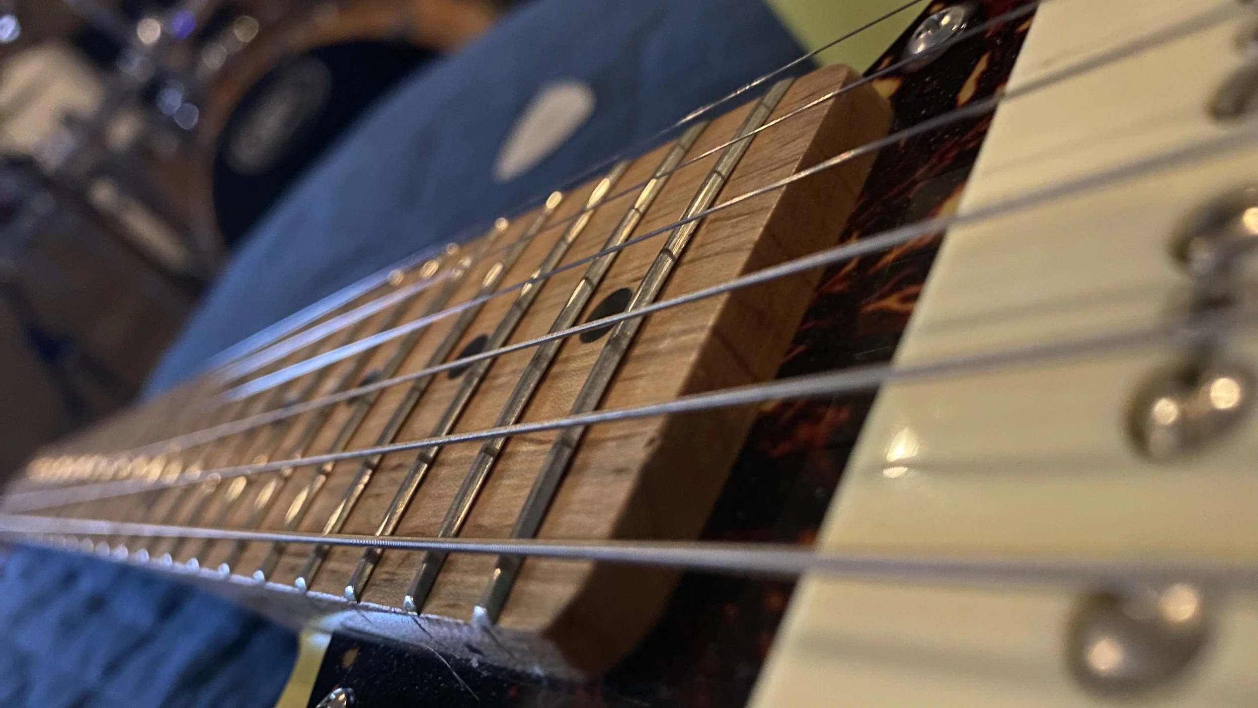 Close-up of a guitar neck showing metal frets, wooden fretboard, and wires across the strings, with a blurred background.