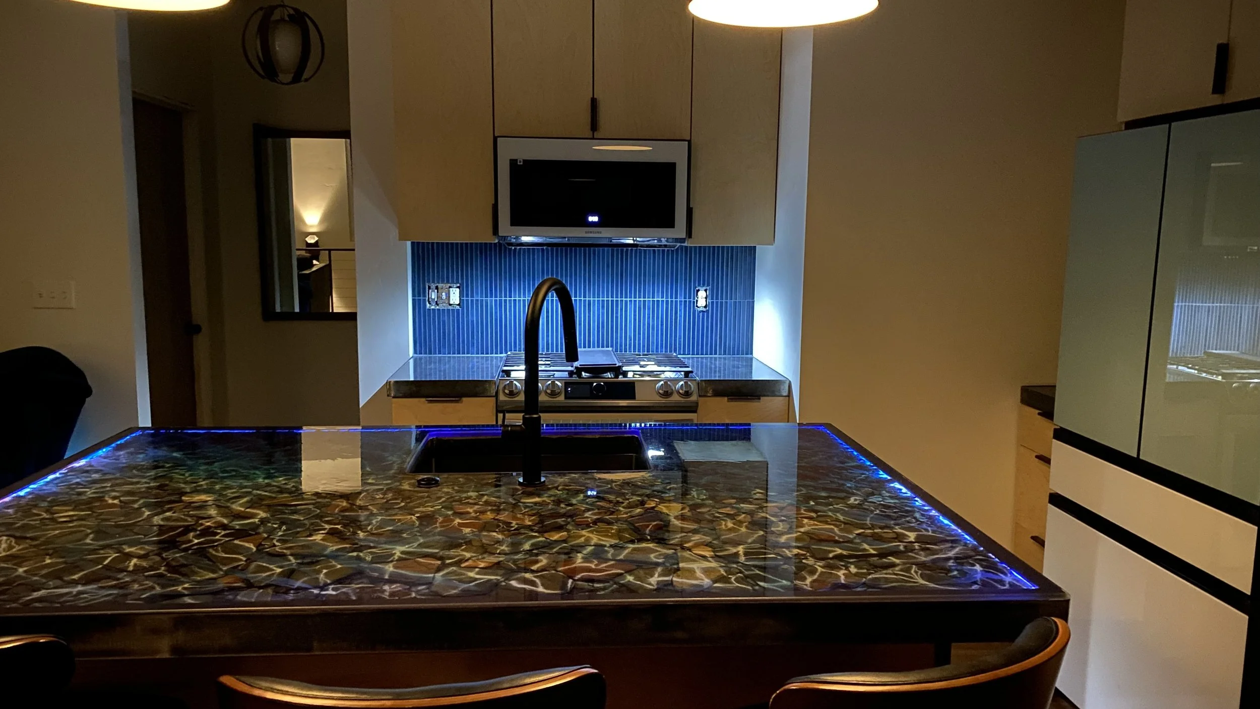 Modern kitchen with a mosaic tile countertop, black faucet, blue tiled backsplash, microwave oven, stovetop, and white cabinetry, illuminated by hanging pendant lights.