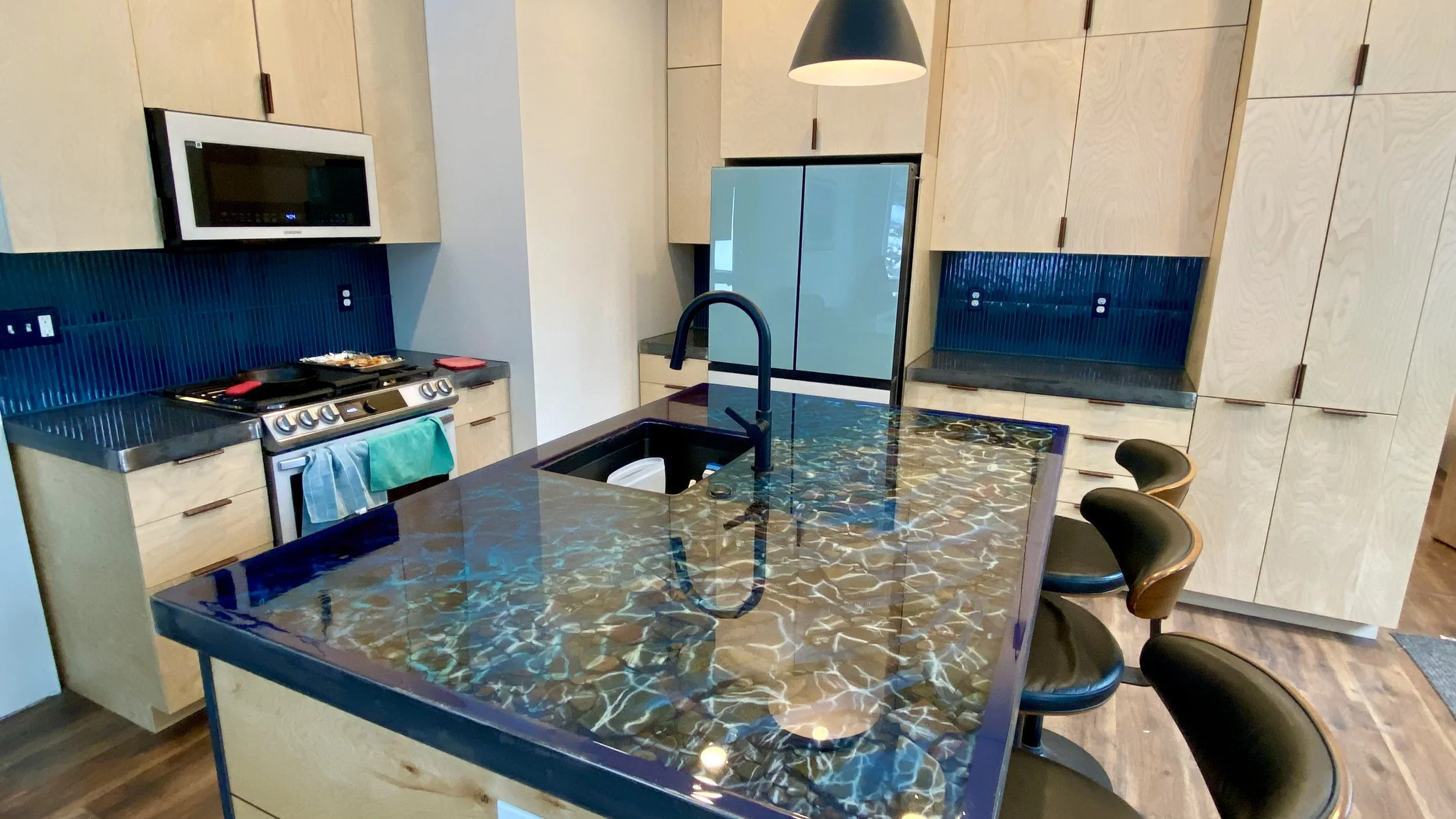 Modern kitchen with light wood cabinets, a black and blue tiled backsplash, a black stovetop, microwave, and a black granite island with a sink and chairs.
