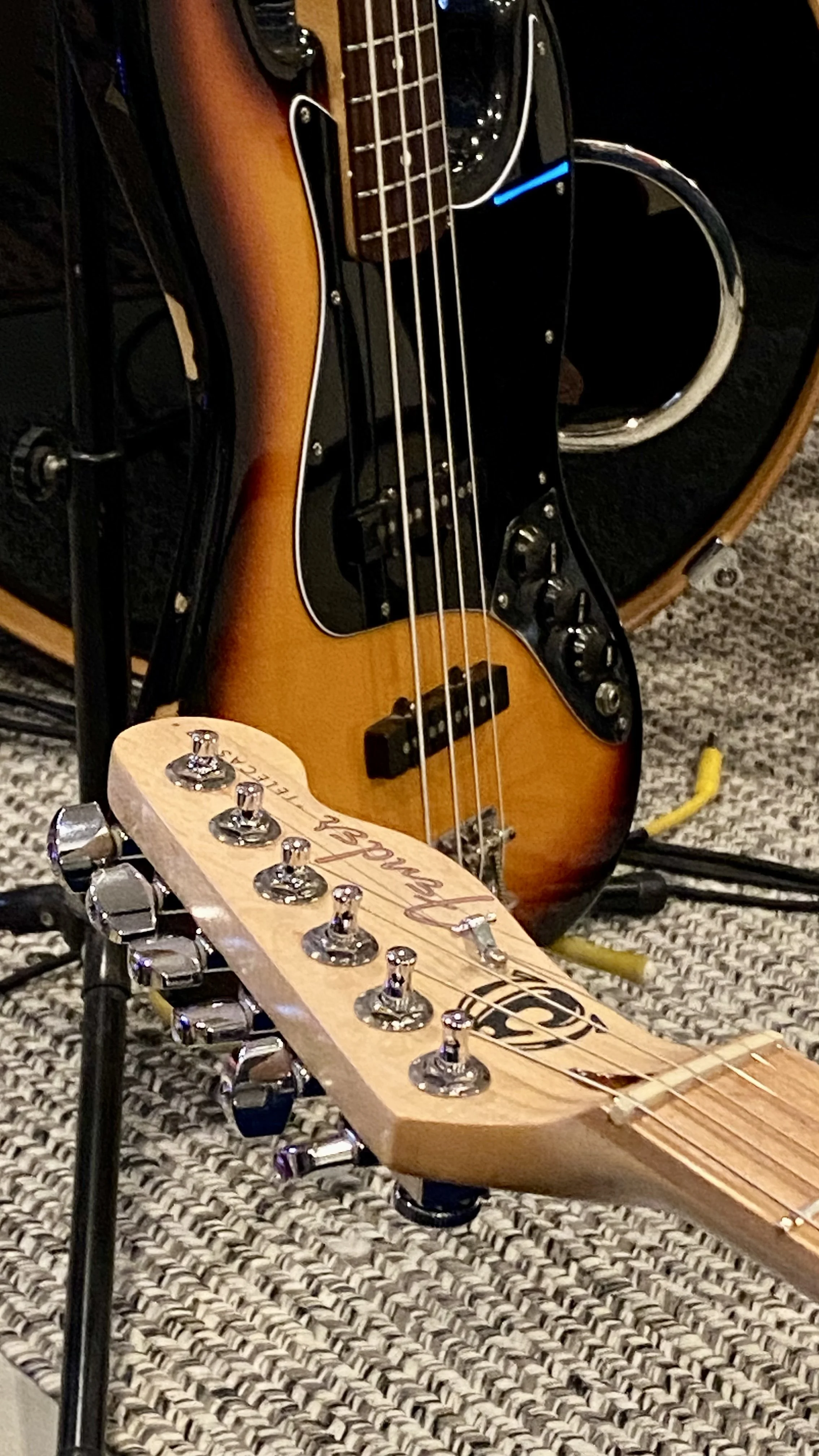 Close-up of a bass guitar and another guitar headstock resting on a textured carpet, with cables and a bass amplifier in the background.
