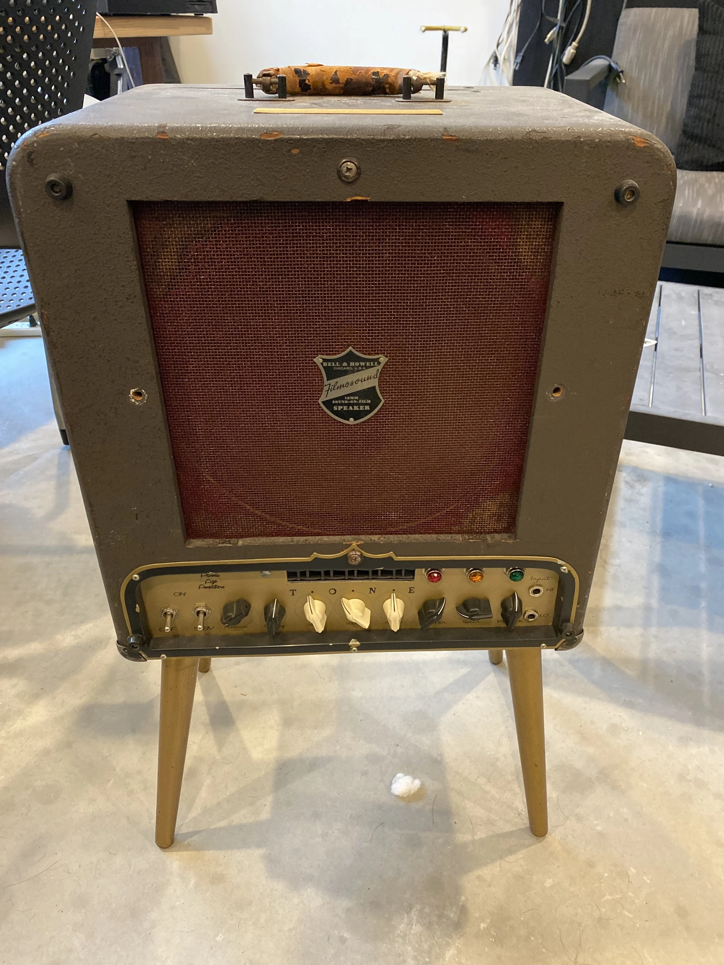 Vintage small brown guitar amplifier with a red grille, standing on four wooden legs, with control knobs and switches on the front panel.
