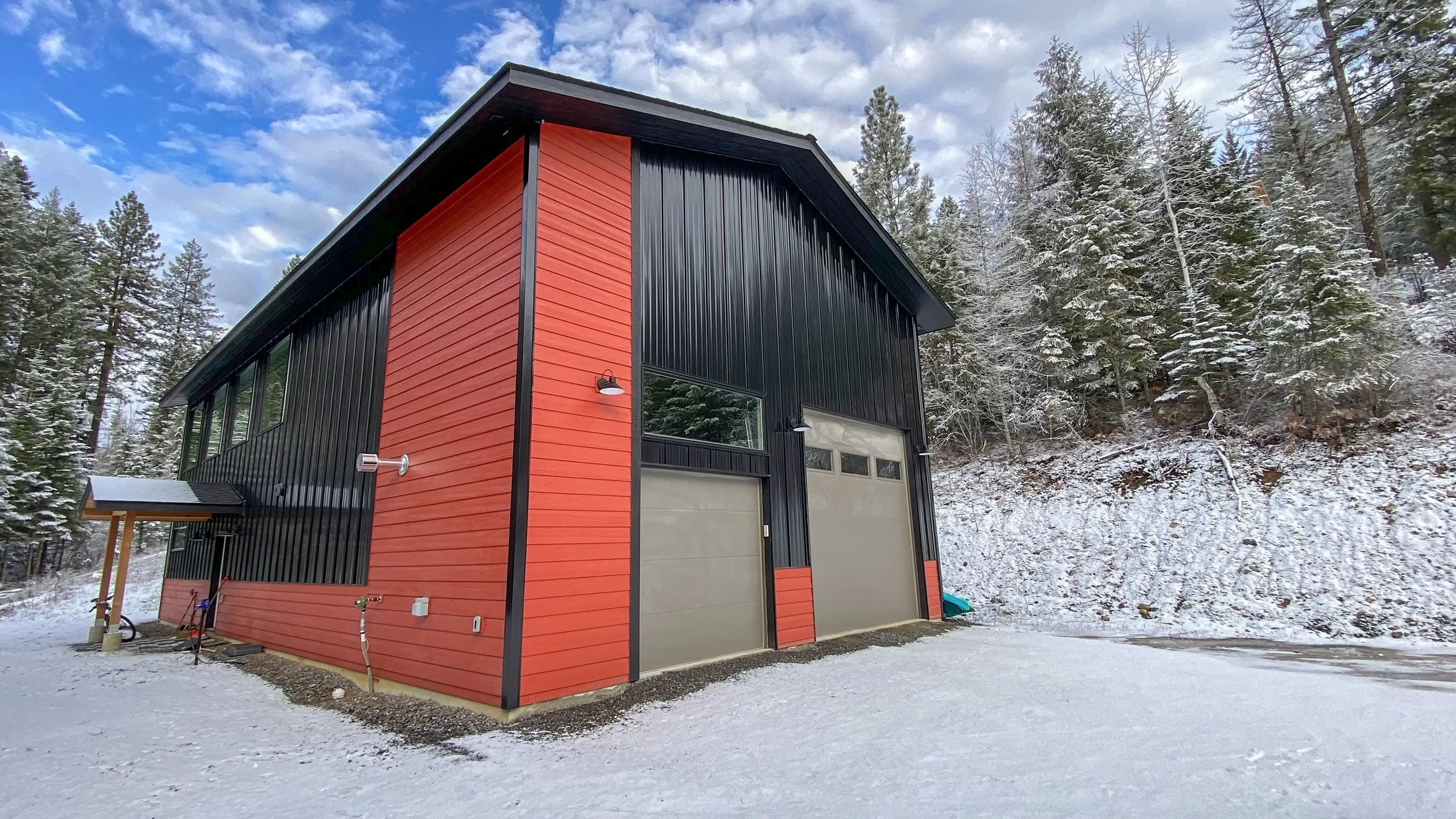 A modern two-story garage with black and red siding, surrounded by snow-covered ground and pine trees, under a partly cloudy sky.