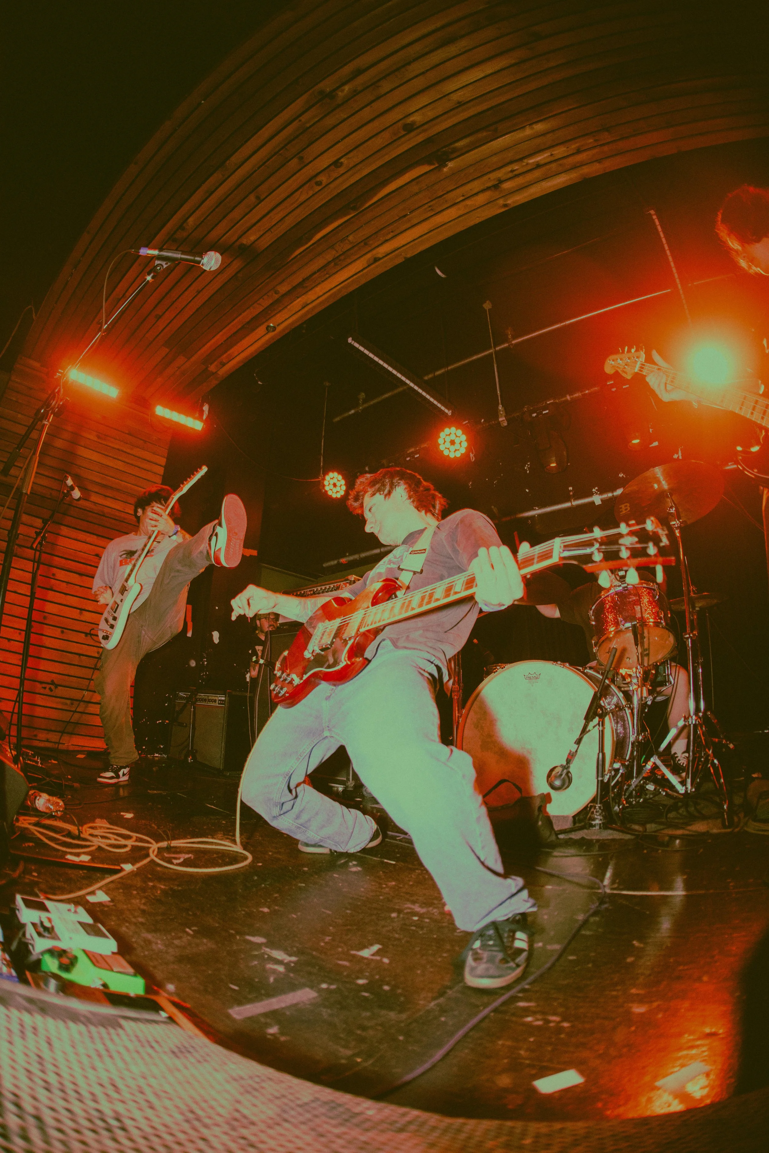 Band performing live on stage with guitars and drums, red and orange stage lighting.