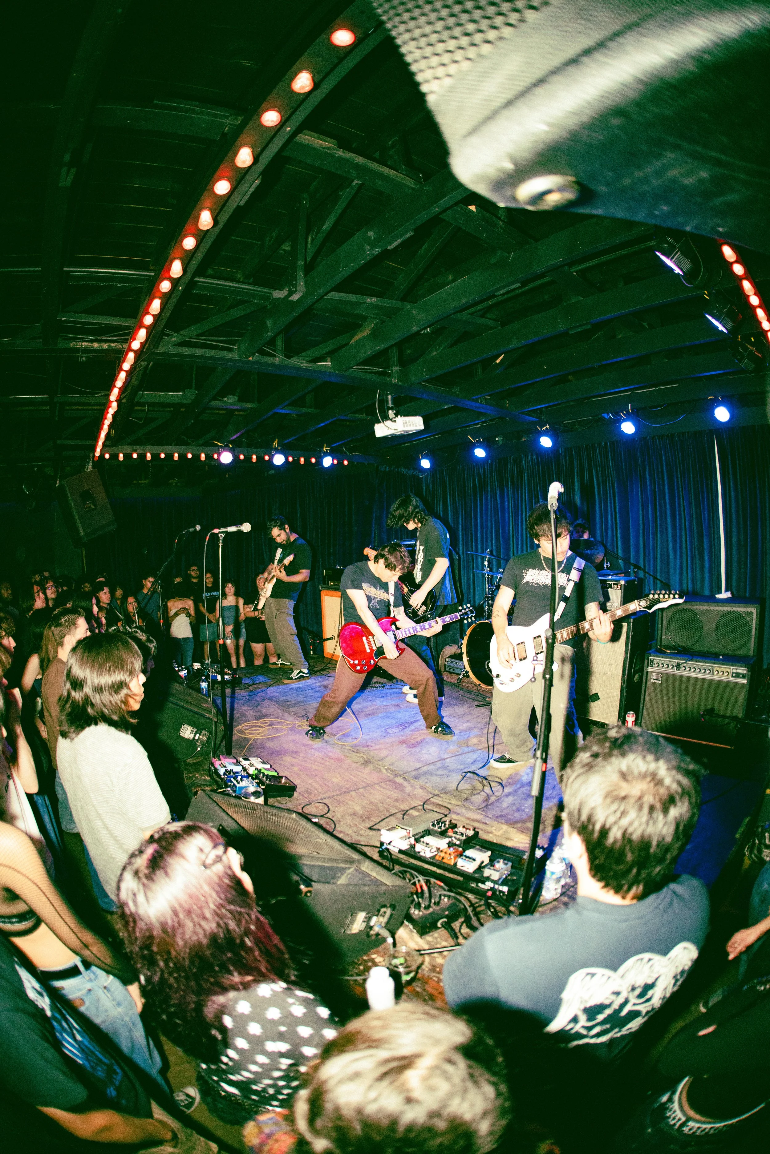 A band performs on stage in a dark venue with blue curtains. The lead singer is playing a red guitar, and other band members are playing guitars. The audience watches from in front of and to the sides of the stage, some standing and some seated. The 
