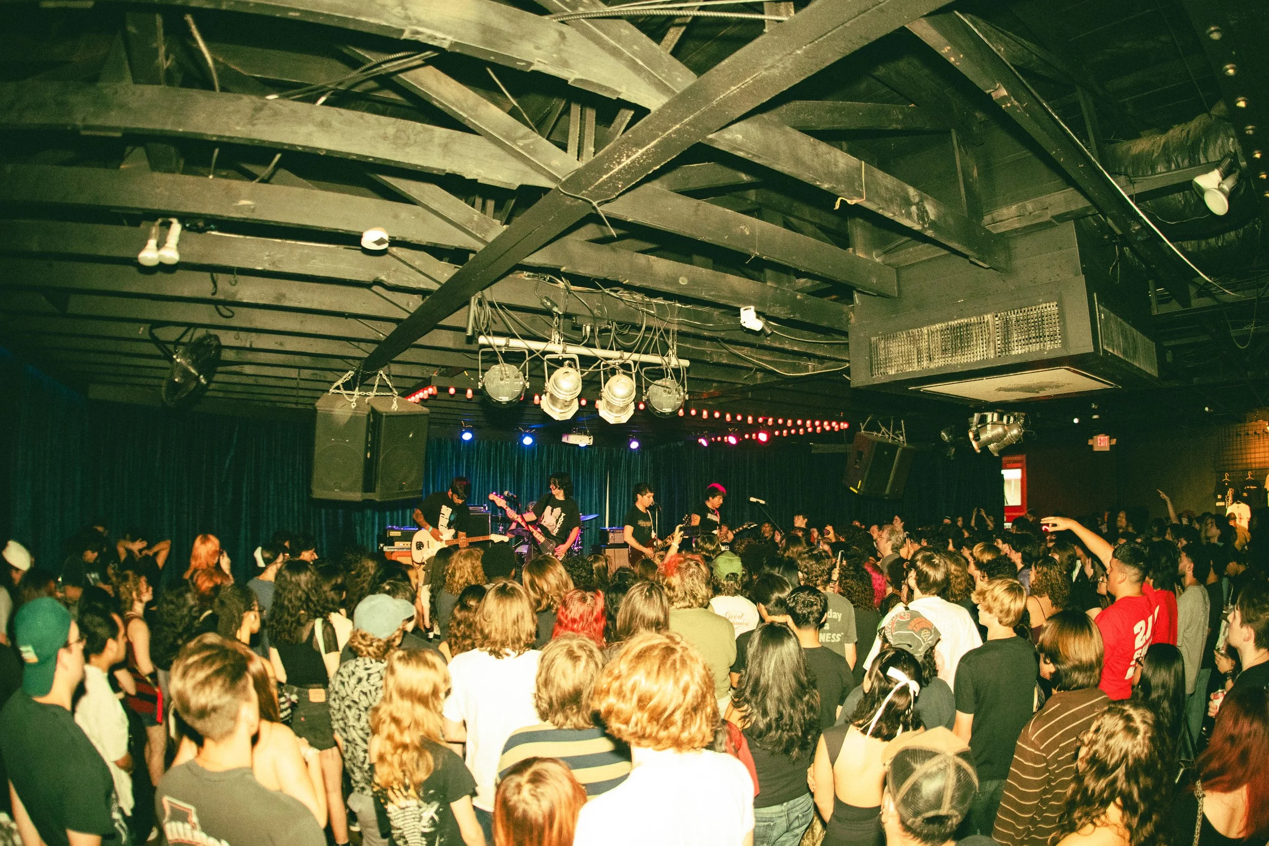 Crowd watching a live band perform on stage in an indoor venue with black curtains and colored lights.