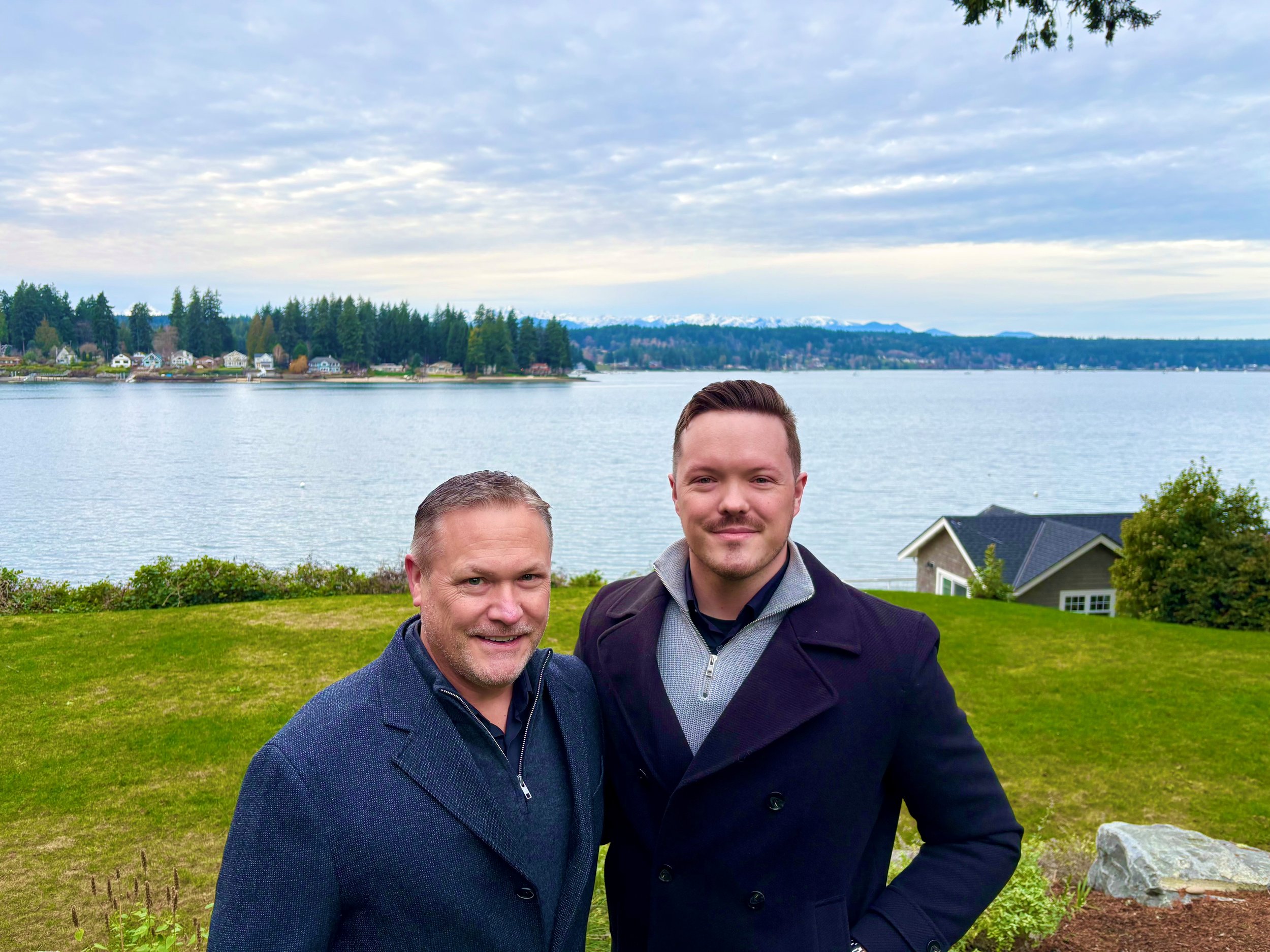 Two men standing outdoors near Liberty Bay, Poulsbo, with houses and trees in the background, overcast sky.