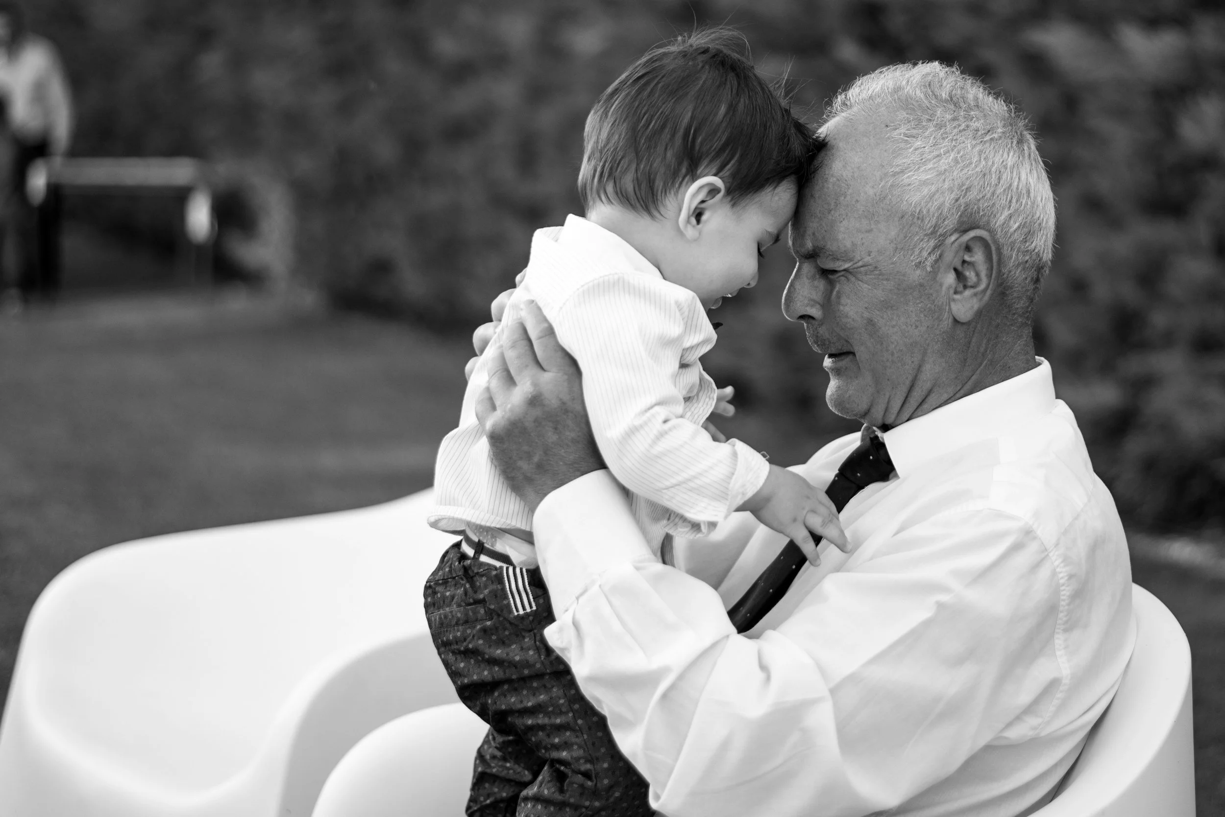 An elderly man holding a young boy, both with their foreheads touching, smiling and sharing a tender moment outdoors.