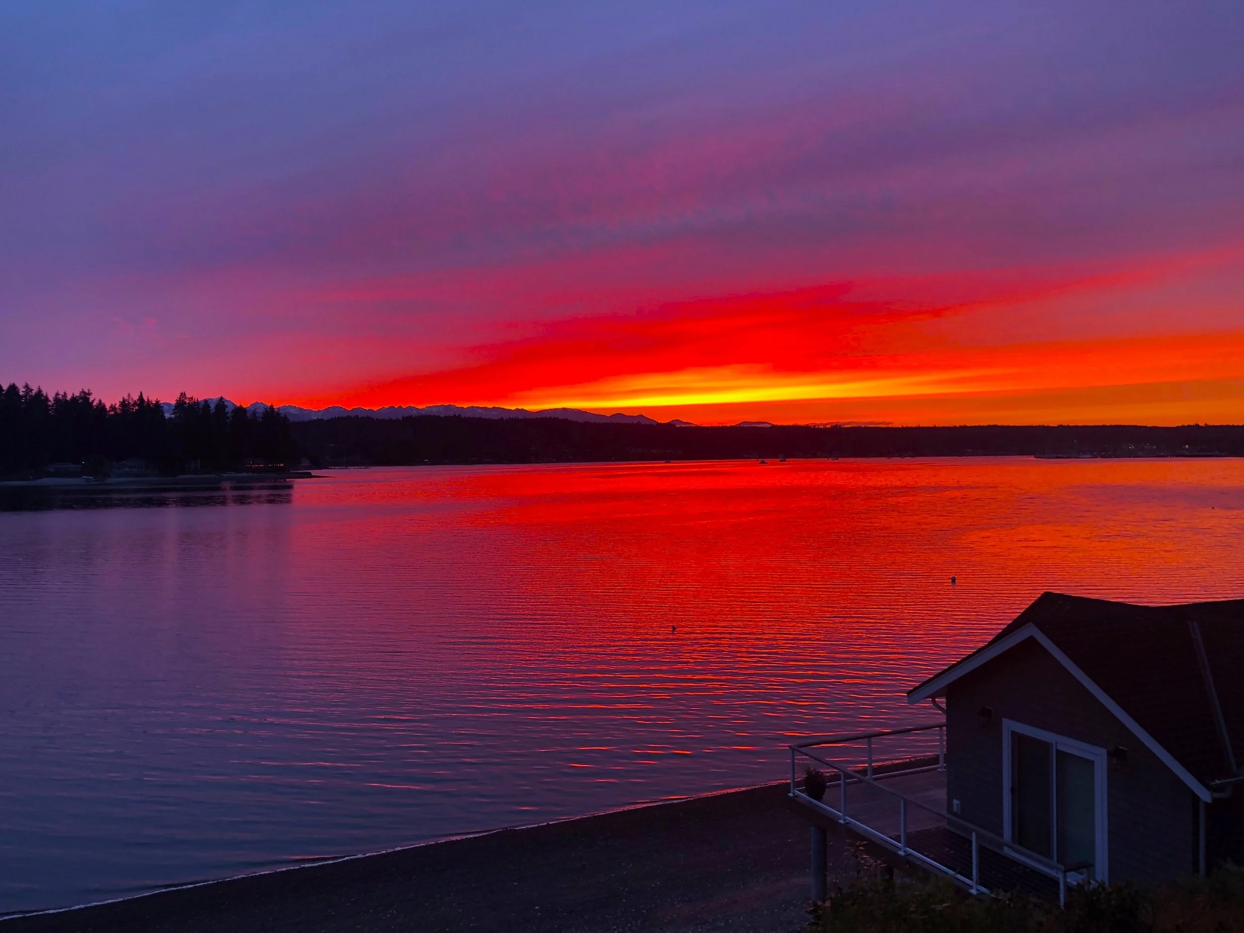 Serene sunset over a calm body of water with colorful sky, distant mountains, trees, and a small house near the shoreline.