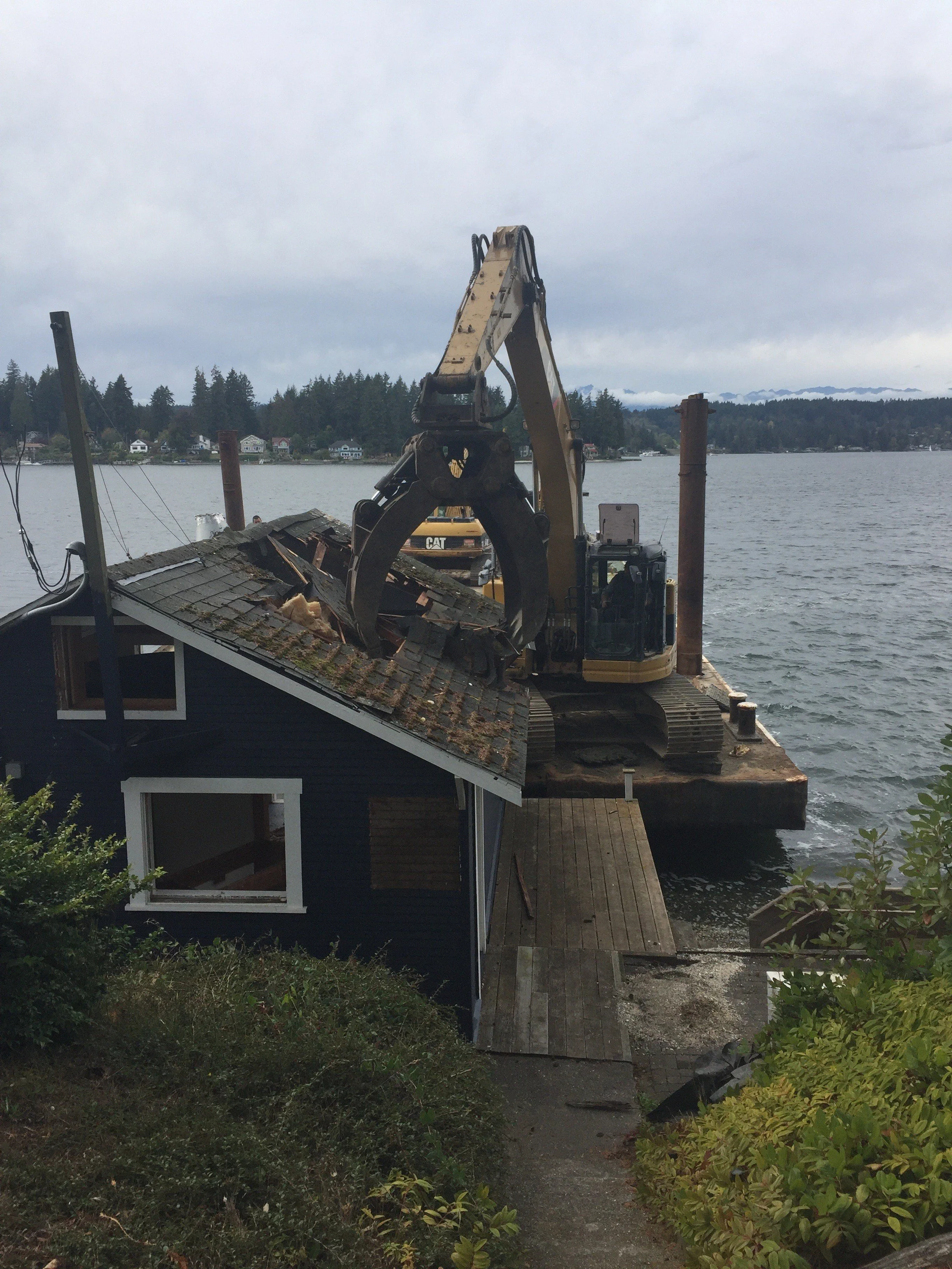 A house being demolished by an excavator near a body of water with houses across the lake and trees in the background.