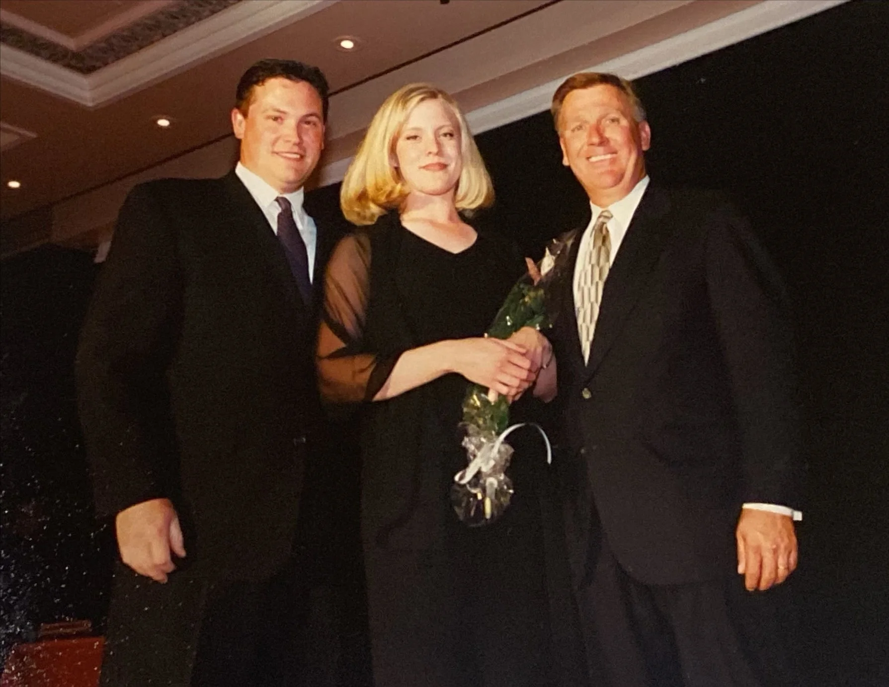 Three people dressed in formal attire standing together, one woman holding a rose, in a dimly lit indoor environment.