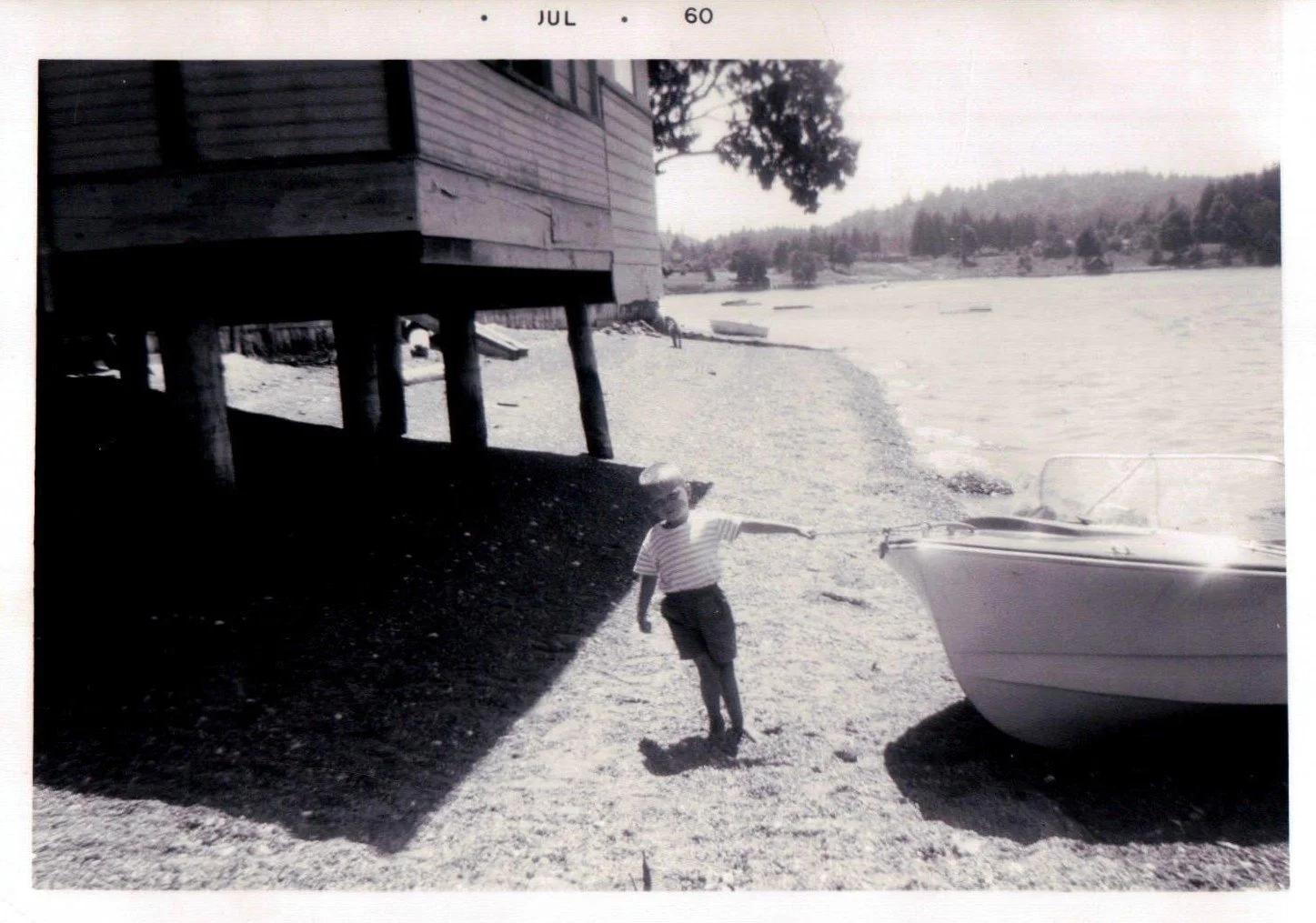 A black and white photograph of a boy in striped shirt and shorts at a lakeside, pointing towards a boat nearby. The boy is standing on sandy ground near a wooden house on stilts, with a tree and distant hills in the background.