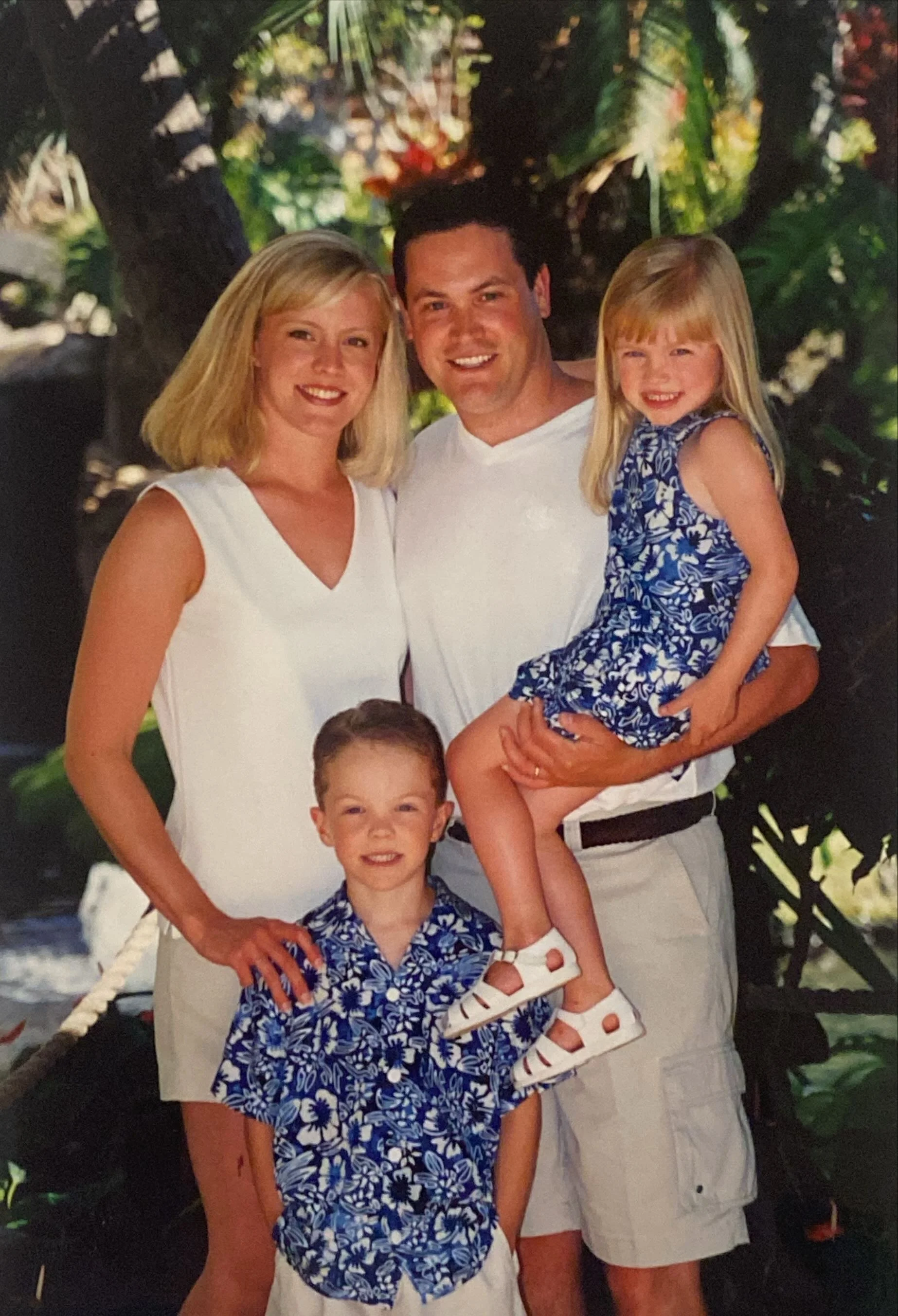 A family of four posing outdoors in front of lush greenery. The woman and man are smiling, with the woman wearing a white dress and the man in a white shirt. The little girl is sitting on the man's arm, wearing a blue floral dress and white sandals, while the young boy stands in front, wearing a matching blue floral shirt.