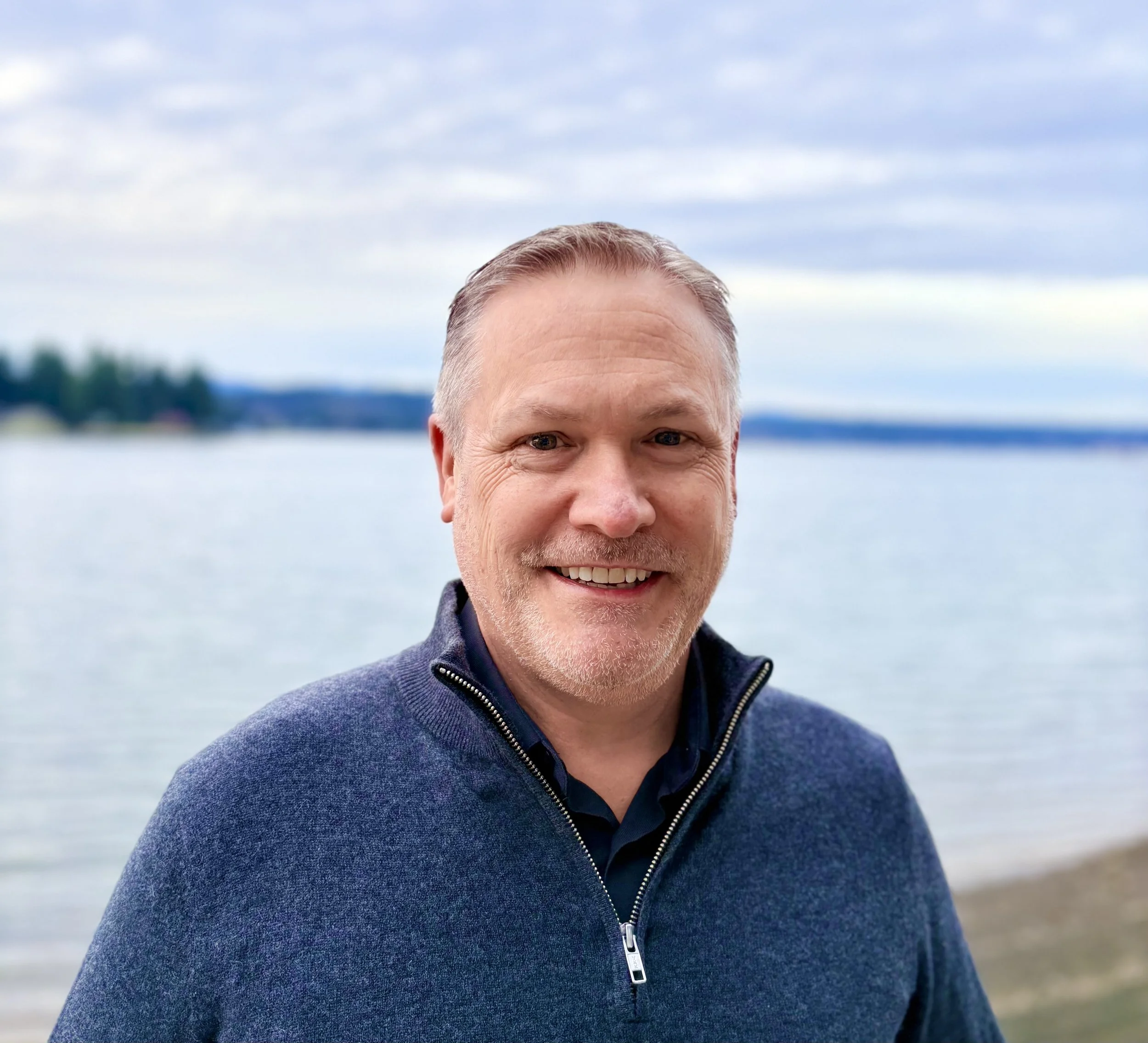 A middle-aged man with short gray hair and a beard, smiling, standing by a body of water with a cloudy sky in the background.