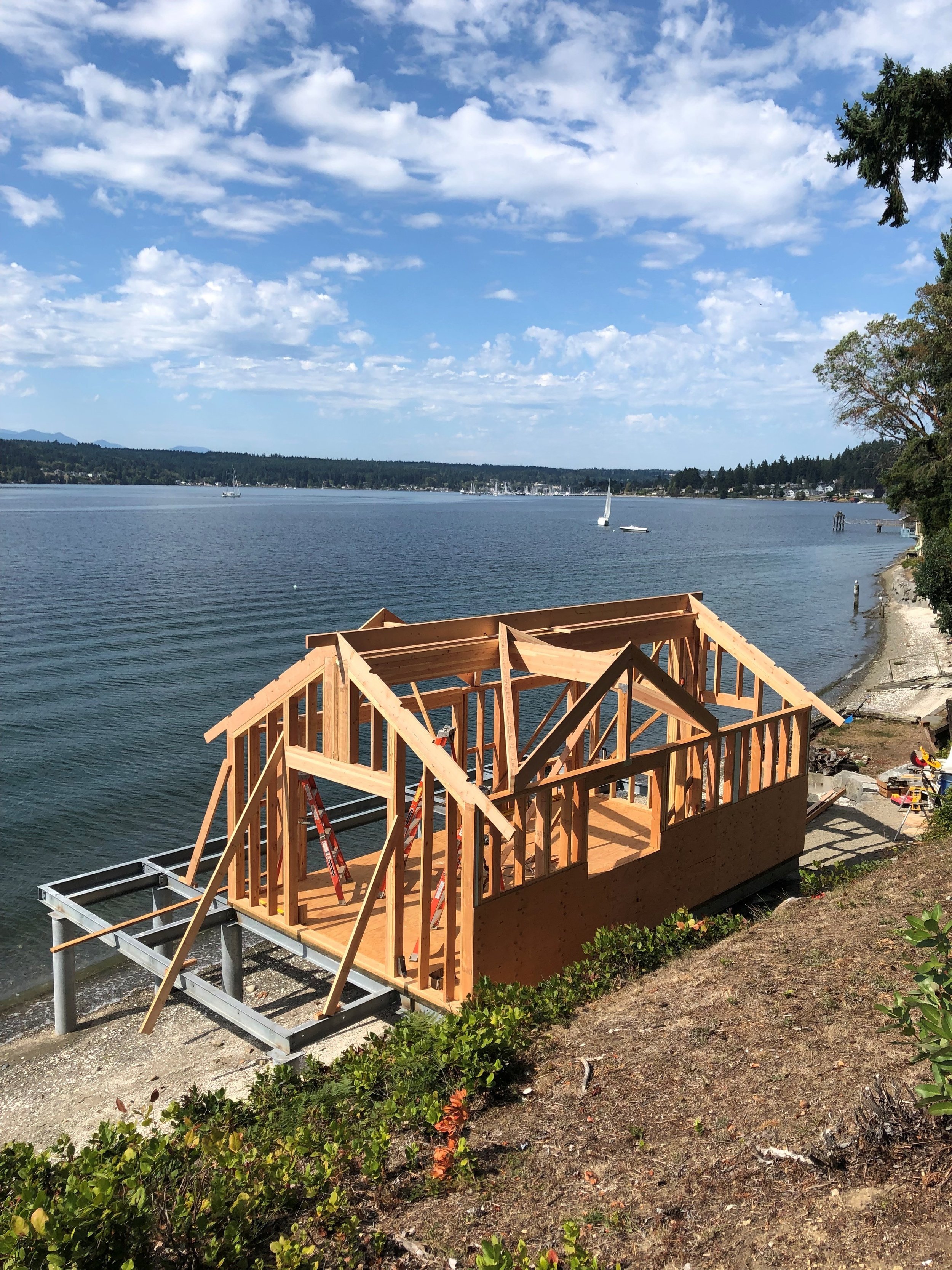 Wooden house frame under construction on a lakeside with sailboats, trees, and a partly cloudy sky in the background.
