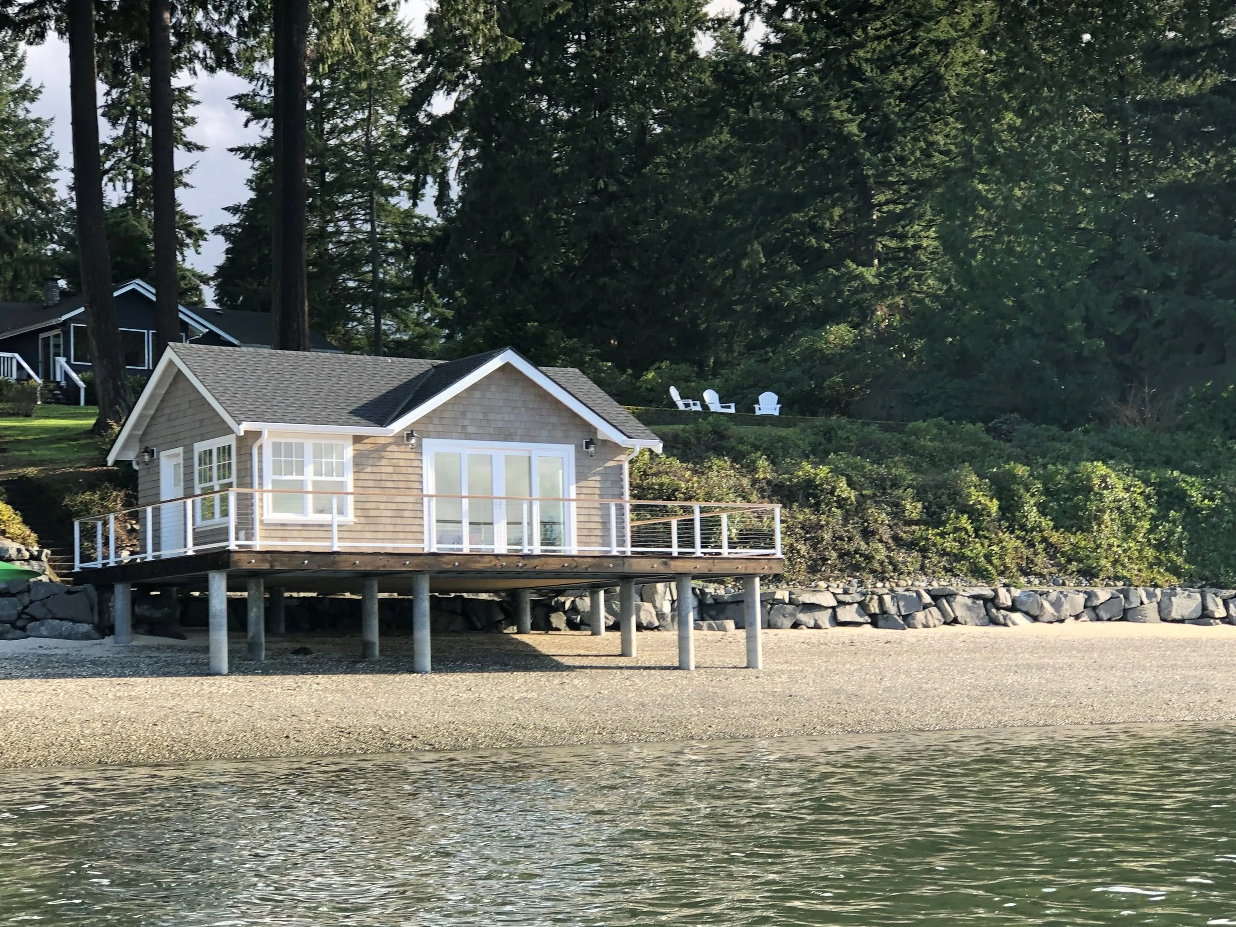 A house on stilts located by the beach, with a deck and sliding glass doors, surrounded by trees and bushes, and a few white chairs on the hillside in the background.