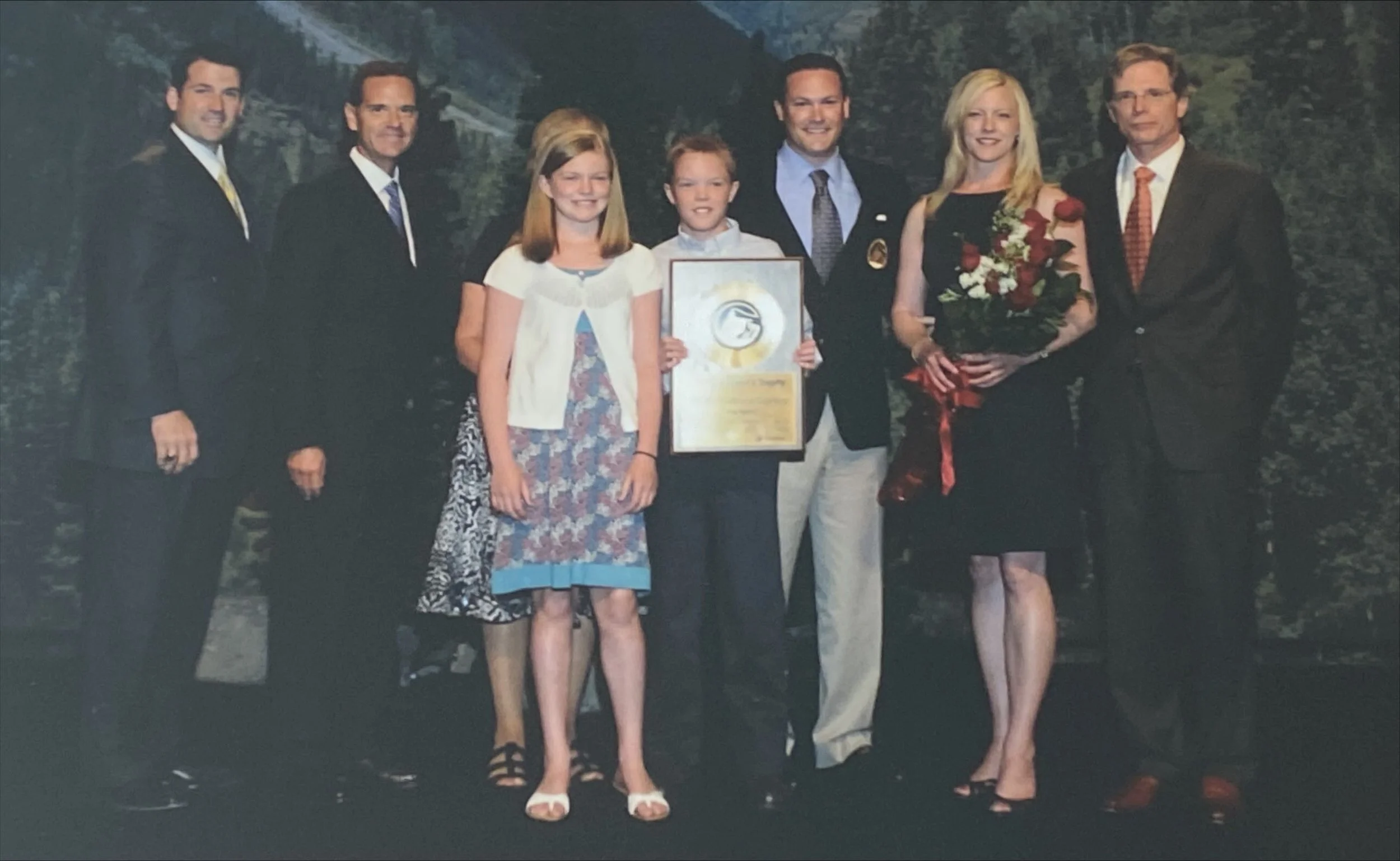 Group of nine people standing outdoors, one girl and boy holding a plaque, a woman holding a bouquet, all dressed in formal attire, with trees and mountains in the background.