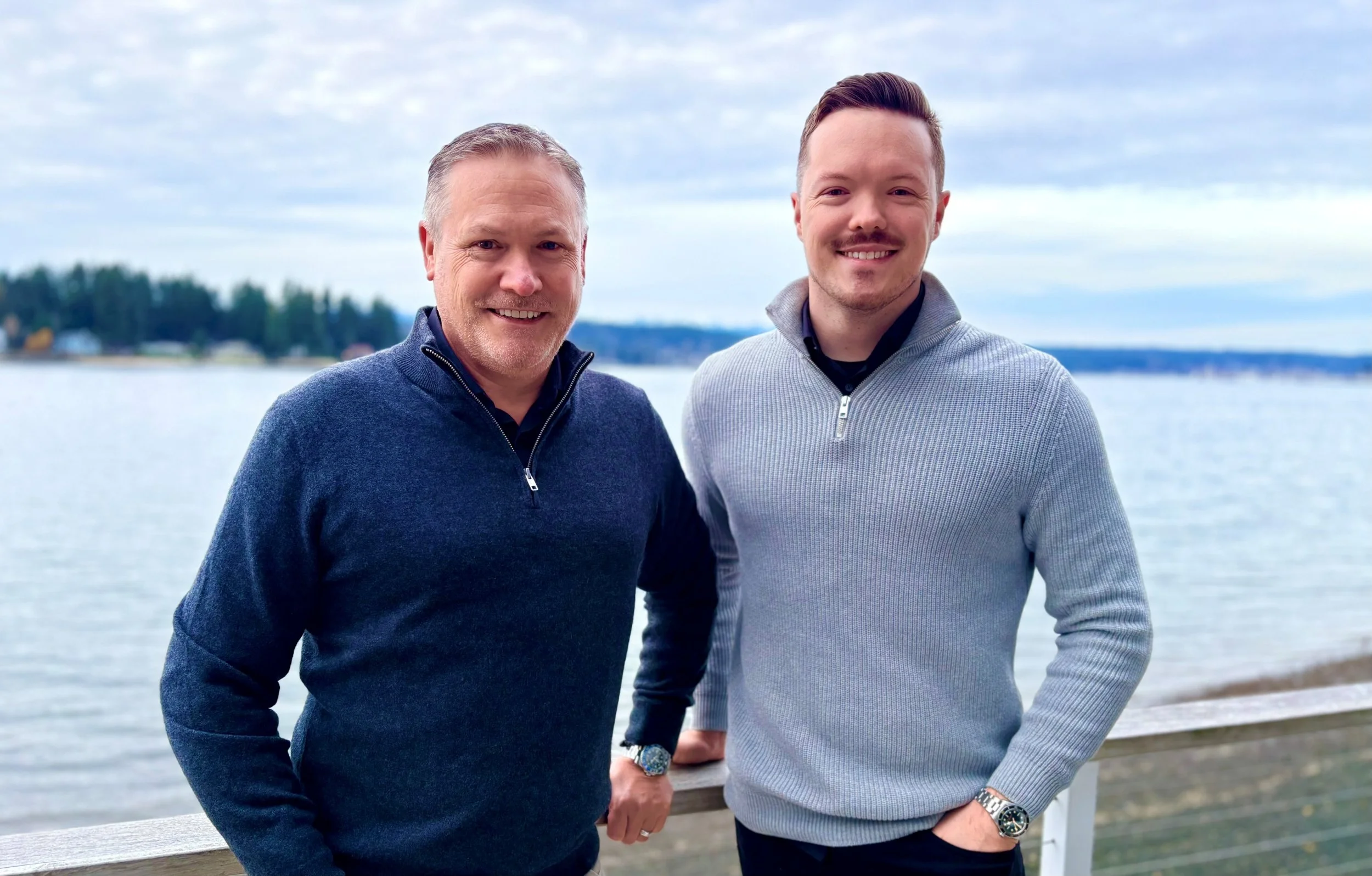 Two men standing outdoors by a body of water with trees and buildings in the background, smiling at the camera.