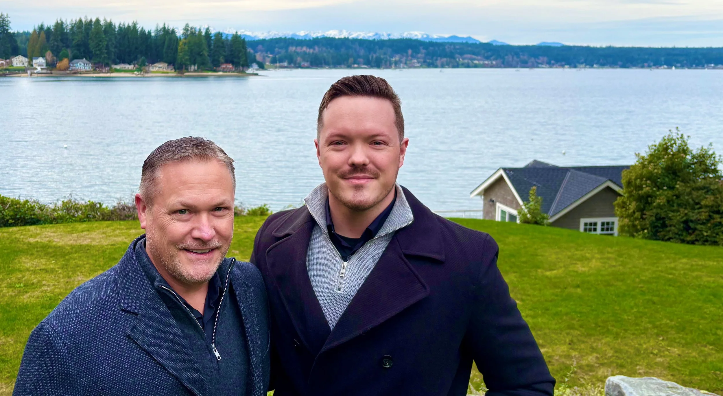 Two men standing outdoors near a body of water with trees, houses, and mountains in the background.