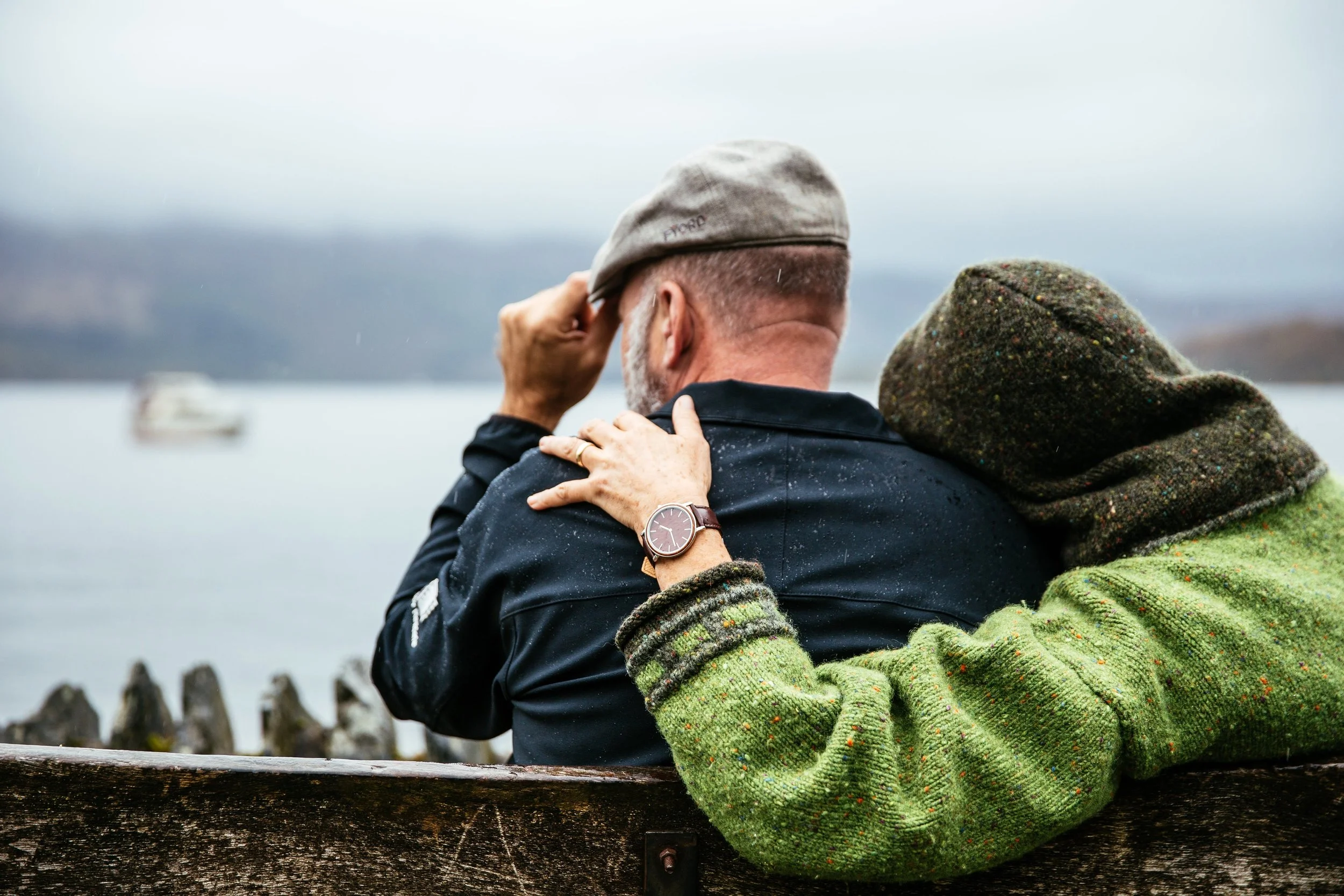 Two men sit on a wooden bench near a body of water, embracing and sharing a moment, with one man wearing a green sweater and a hat, and the other in dark clothing with a watch, under a cloudy sky.