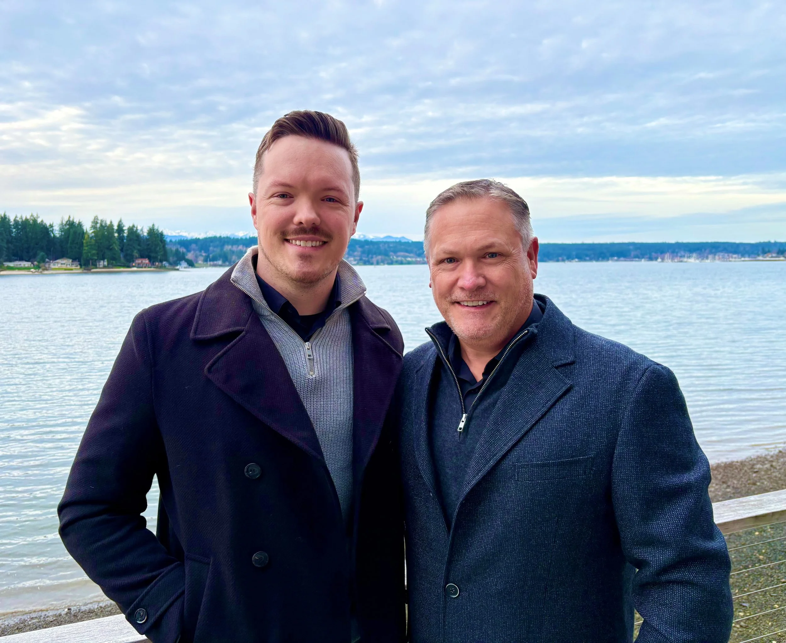 Two men smiling outdoors near a lake with a scenic view of trees, houses, and mountains in the distance, under a partly cloudy sky.