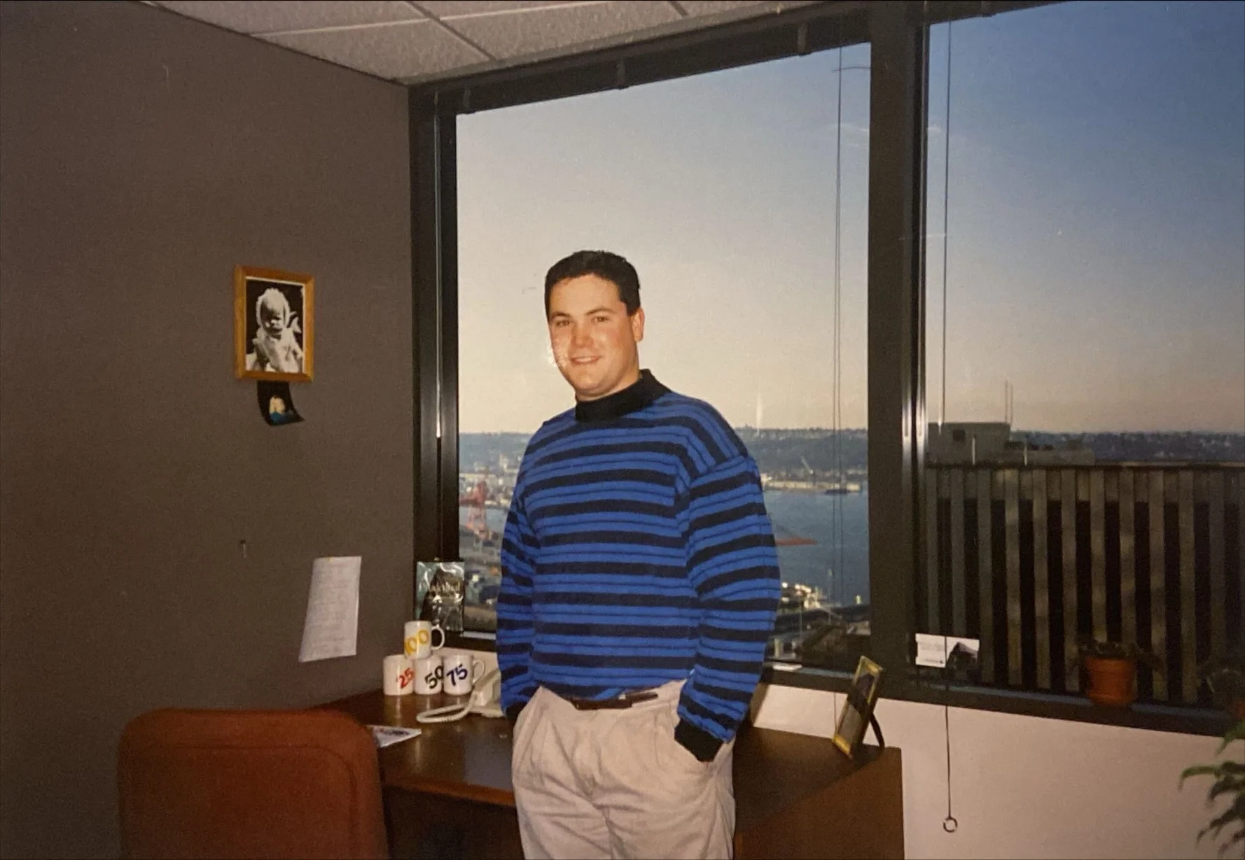 A man wearing a blue and black striped sweater and light-colored pants standing by a desk in an office with a view of a harbor and cityscape through large windows at sunset.