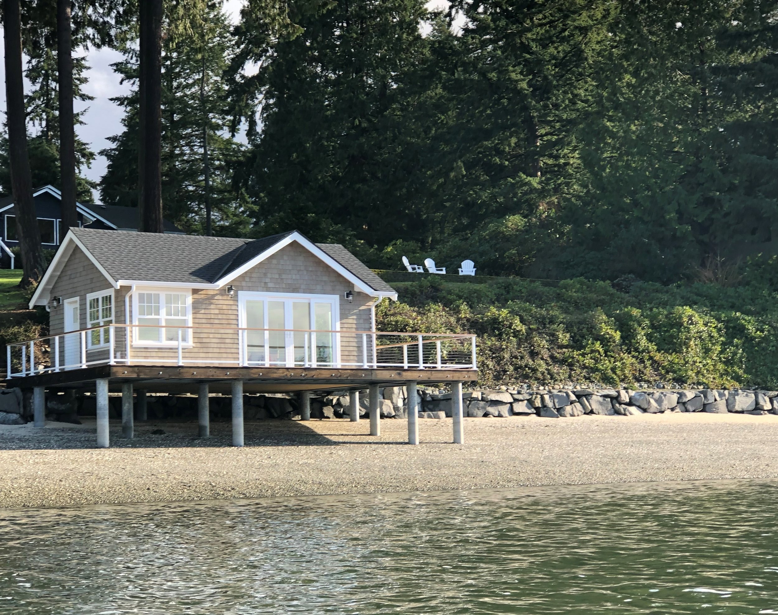A small pier with a house on stilts extending over a body of water. The house has large windows and a deck with white railing. There are three Adirondack chairs on a grassy slope behind the house, surrounded by trees.
