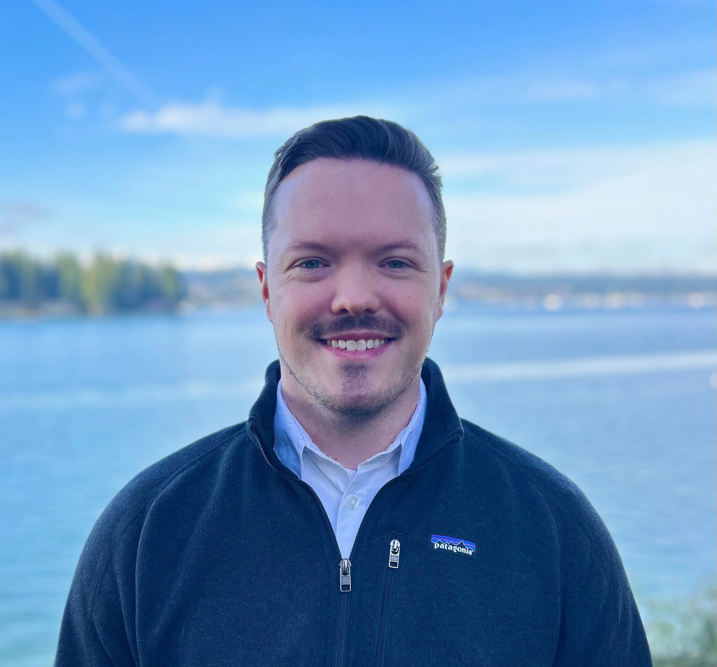 A smiling man outdoors near a body of water during daytime, wearing a black Patagonia fleece jacket over a white collared shirt with a blurred landscape of water and trees in the background.