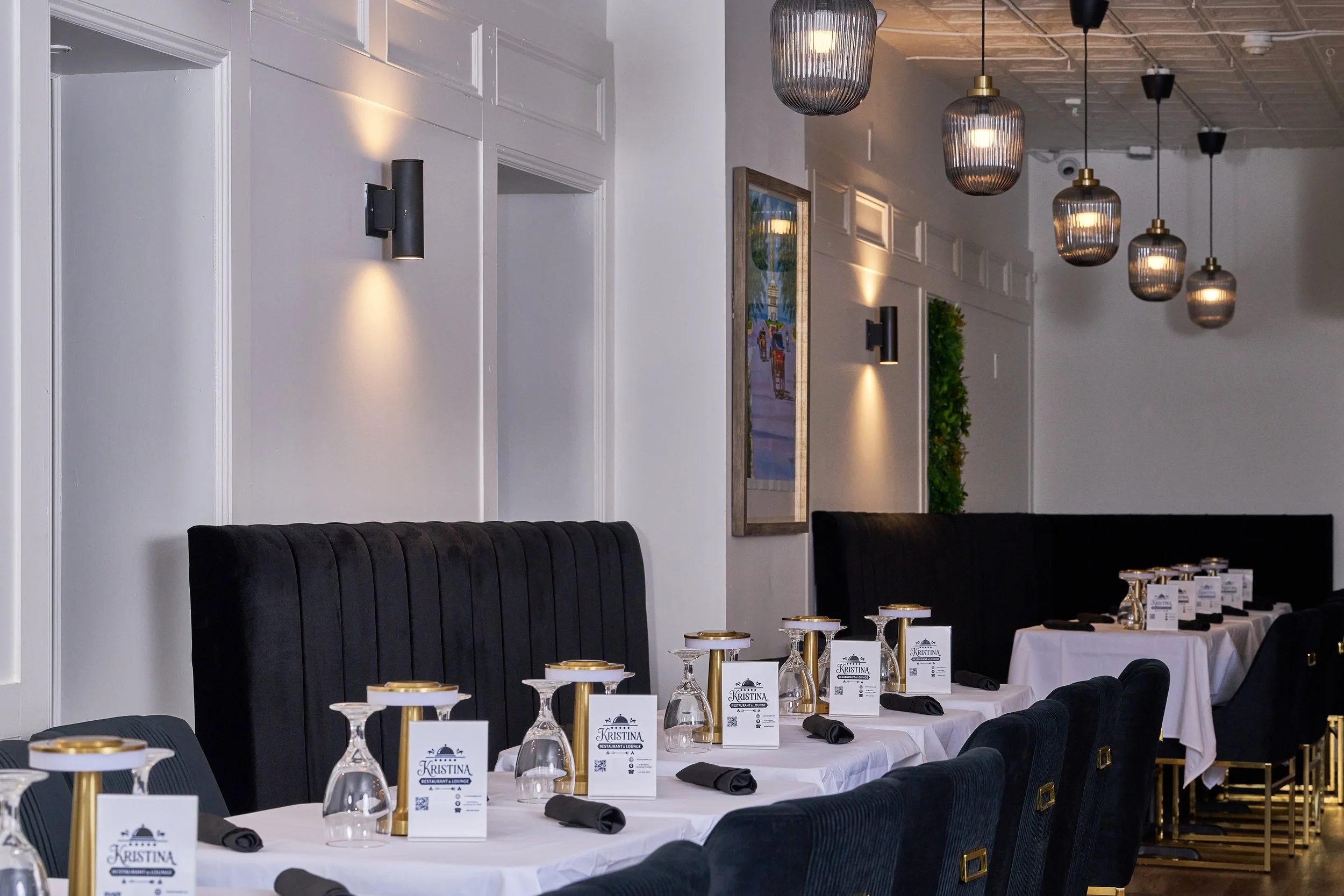 A formal dining room with tables set for a meal, decorated with napkins, glasses, and menus, with black upholstered chairs and wall-mounted lights.