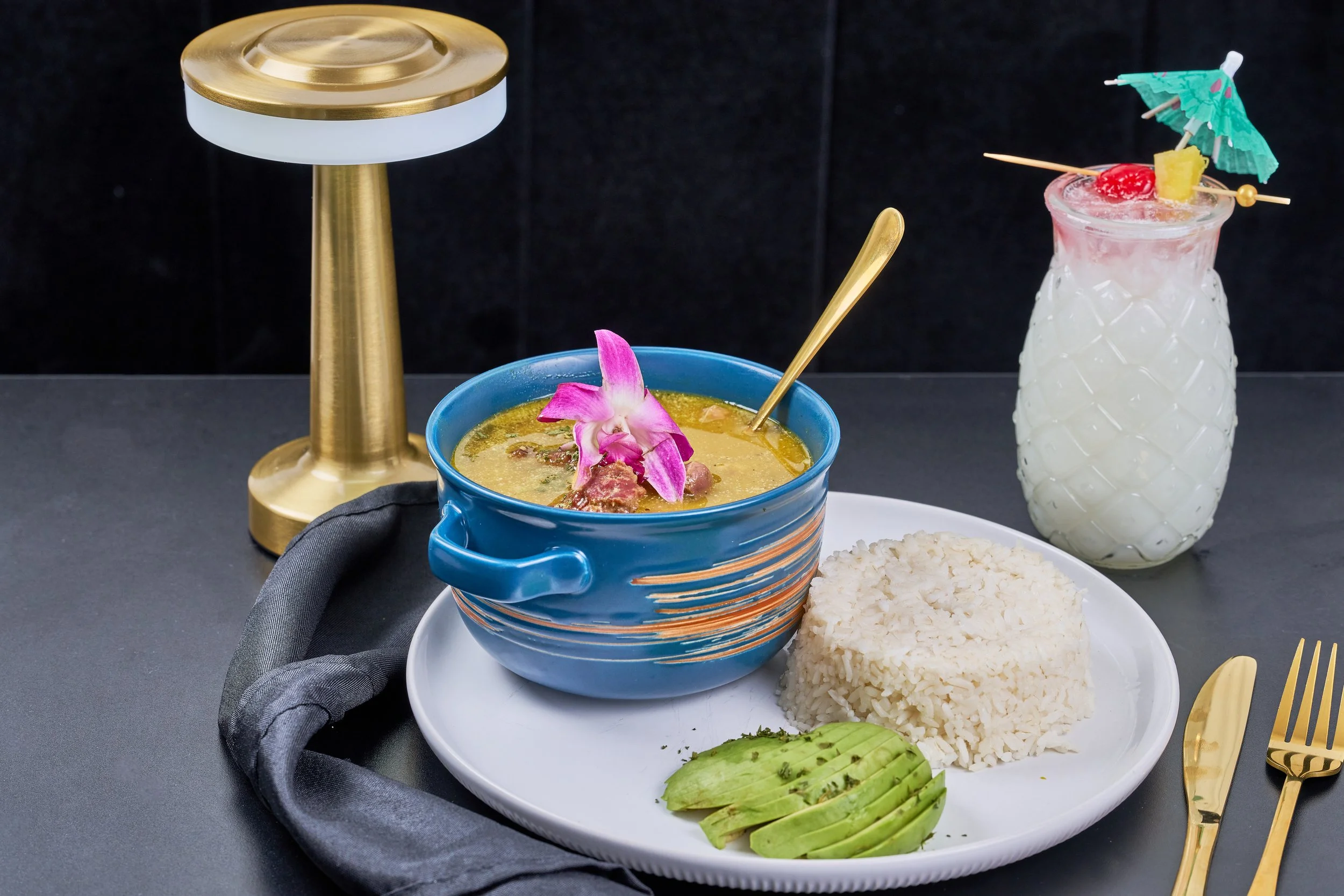 A meal with white rice, green mango slices, and Thai curry garnished with an orchid flower, accompanied by a tropical drink with a small umbrella and fruit garnishes on a black surface with gold utensils.