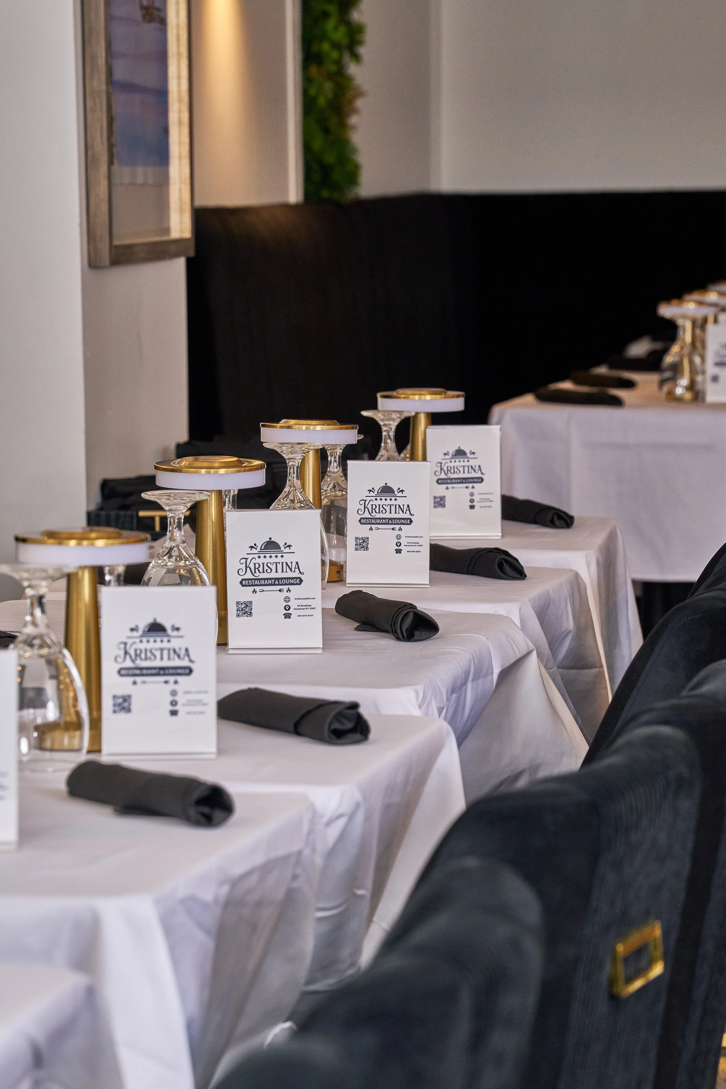 A row of banquet tables set for a formal event, with white tablecloths, black napkins, glassware, and place cards labeled Kristina, in a restaurant or lounge setting.
