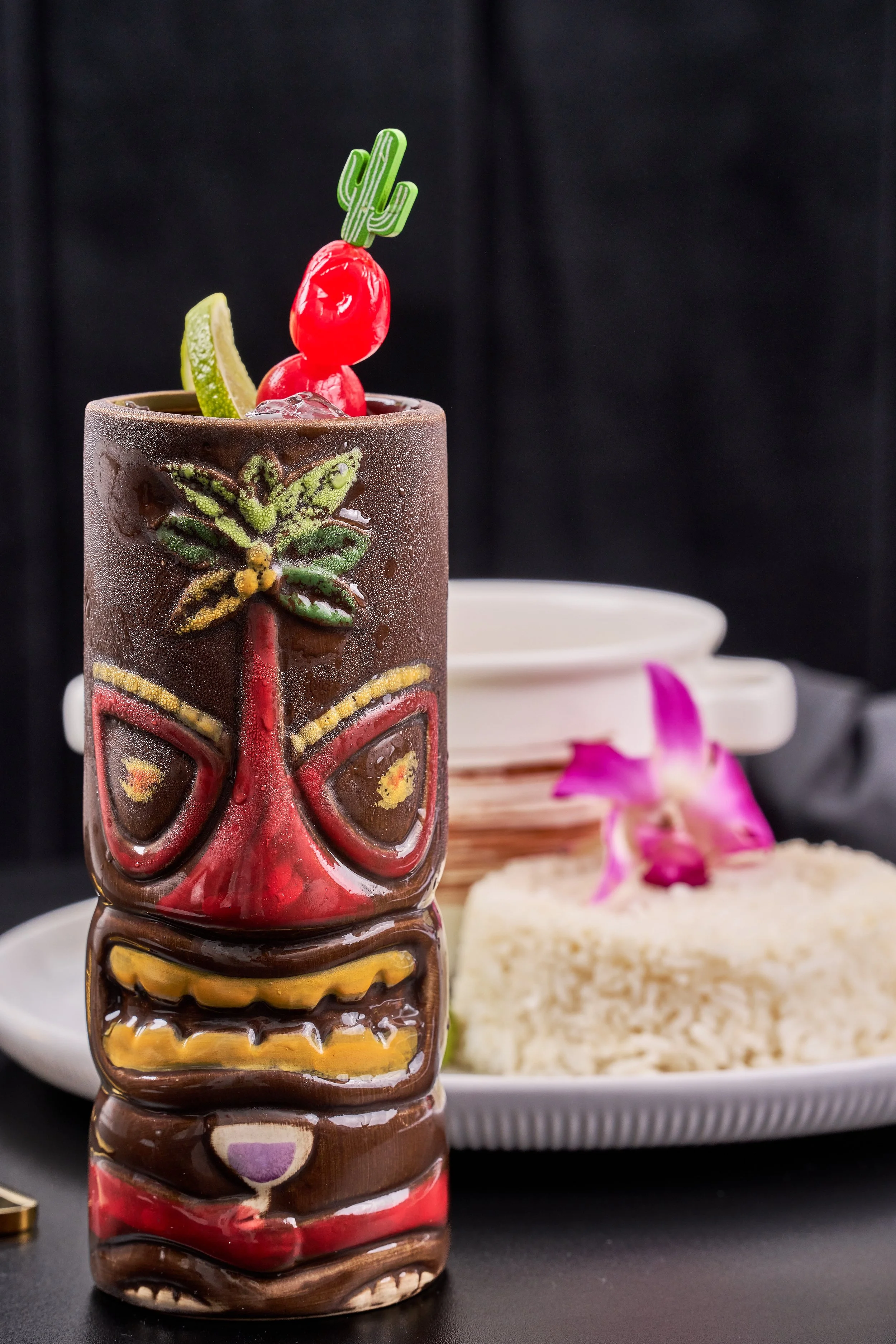 A drink in a tiki mug with a Tiki face design, garnished with a lime wedge, a red cherry, a cherry, and a green cactus-shaped stir stick. In the background, there is a plate with a rice dish and a purple orchid flower.