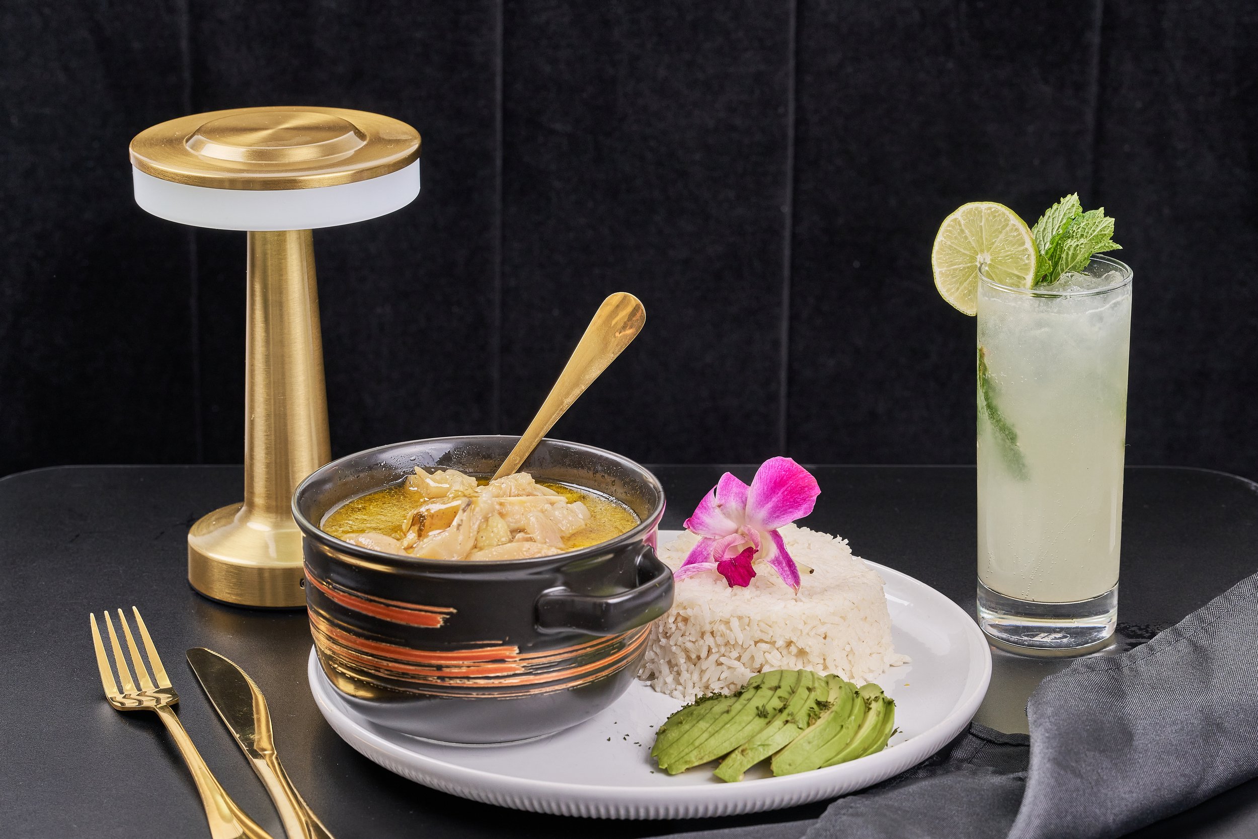 A plate with rice, slices of avocado, and a garnish of purple flower. A black bowl with chicken and sauce, a tall glass of lime and mint drink with ice, a gold-colored lamp, and gold-colored fork and knife on a black table.