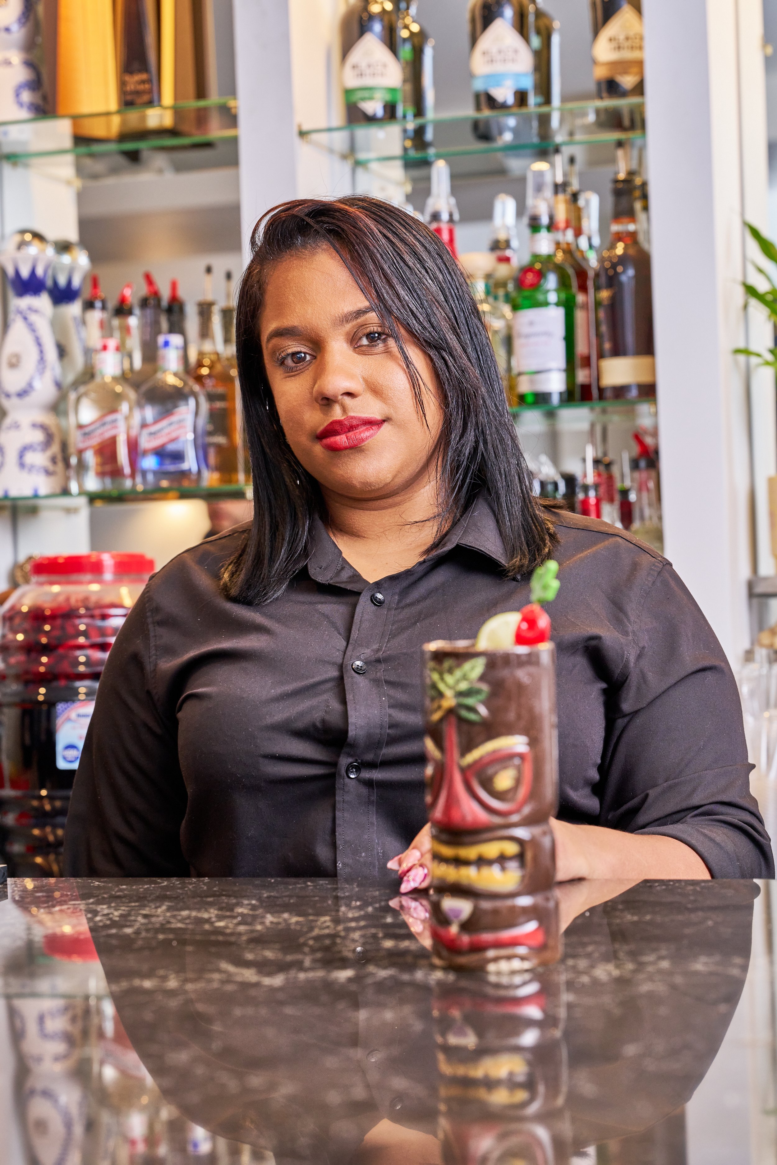 A woman with straight black hair and a black shirt sitting at a bar, with a decorated tiki-style cocktail in front of her.