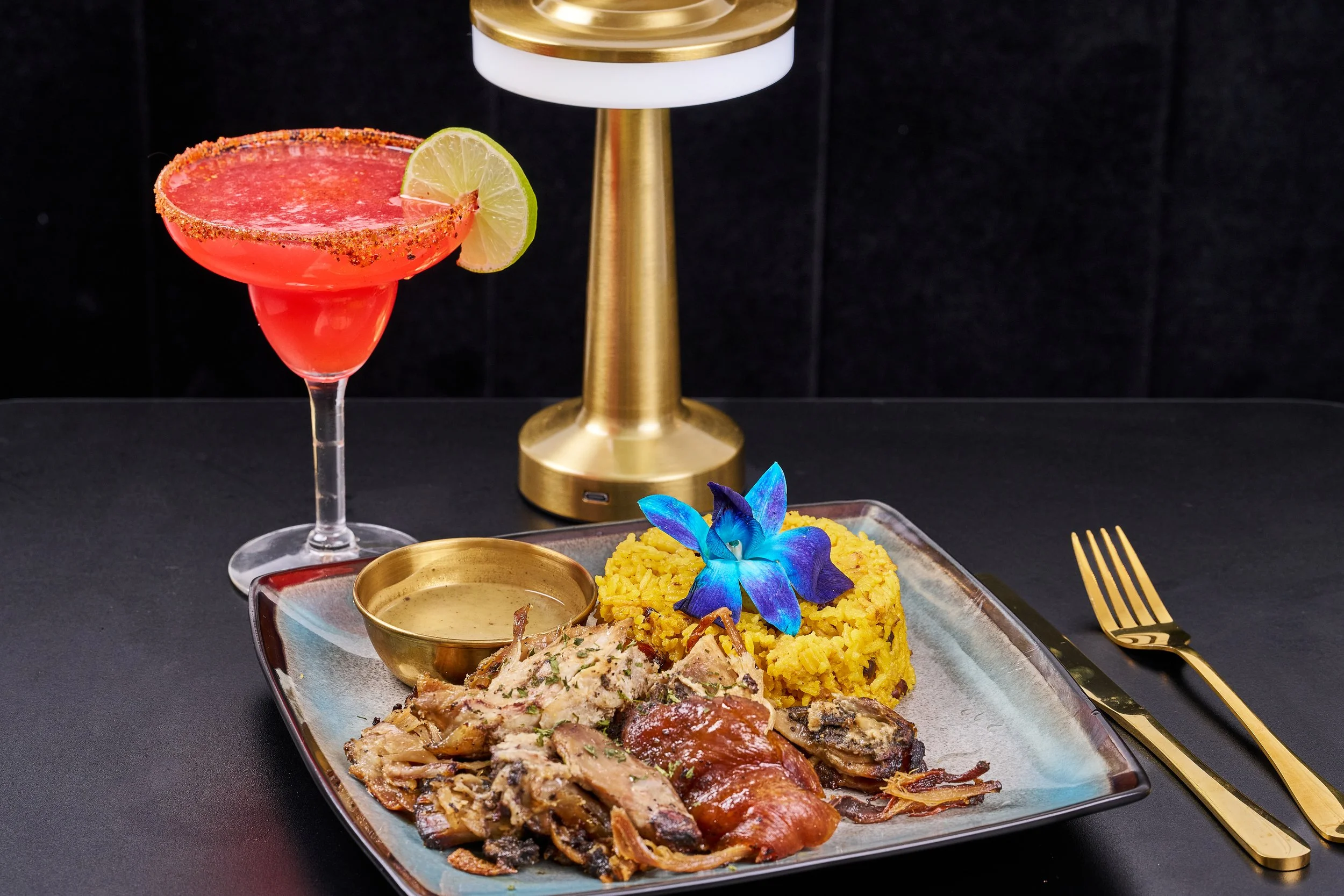 A meal with grilled meat and rice on a plate, a small bowl of sauce, a blue flower for decoration, and a pink cocktail with lime and chili rim on a black table.