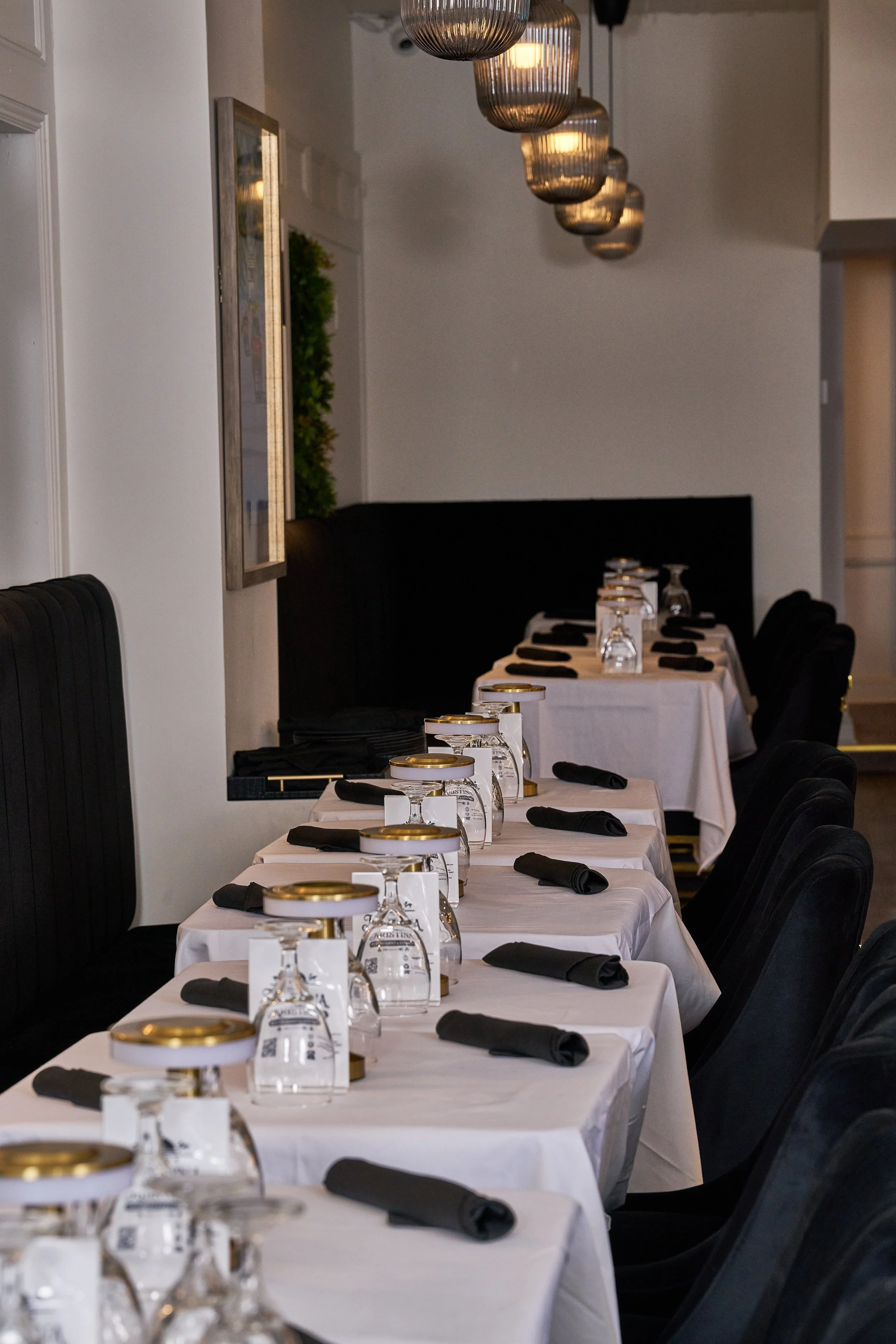 Rows of arranged tables with white tablecloths, black napkins, upside-down wine glasses with gold rims, and small white menu cards, in a restaurant dining area with black chairs and pendant lights.