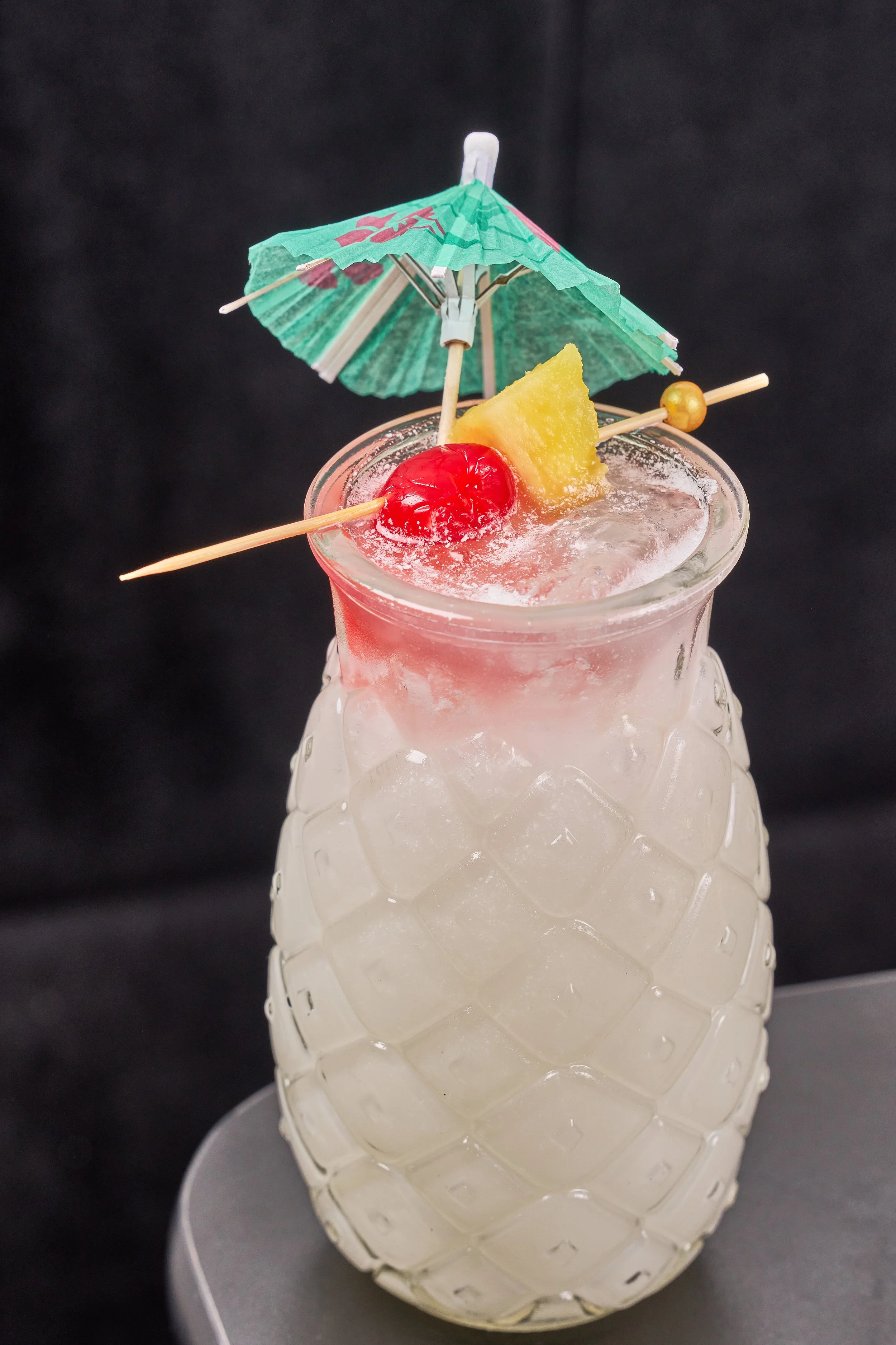 A tropical cocktail in a pineapple-shaped glass with ice, garnished with a cherry, pineapple wedge, and a small paper umbrella.