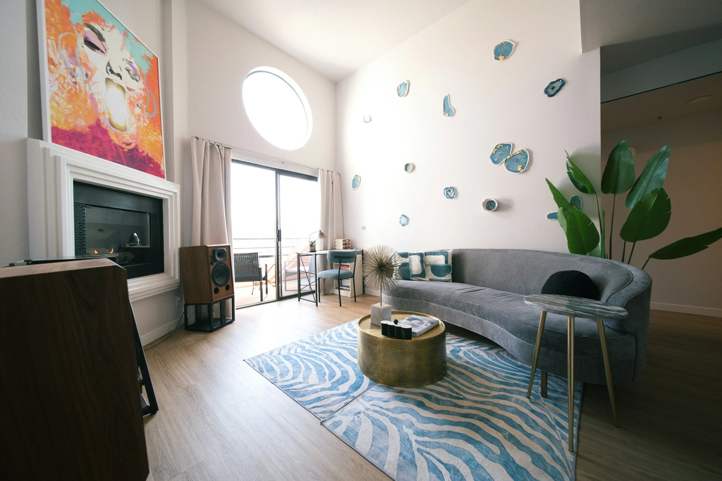 Living room with a gray sofa, blue and white pillows, a gold coffee table, a patterned rug, a large green plant, a fireplace with a colorful art piece above, and a balcony with sliding glass doors and curtains.