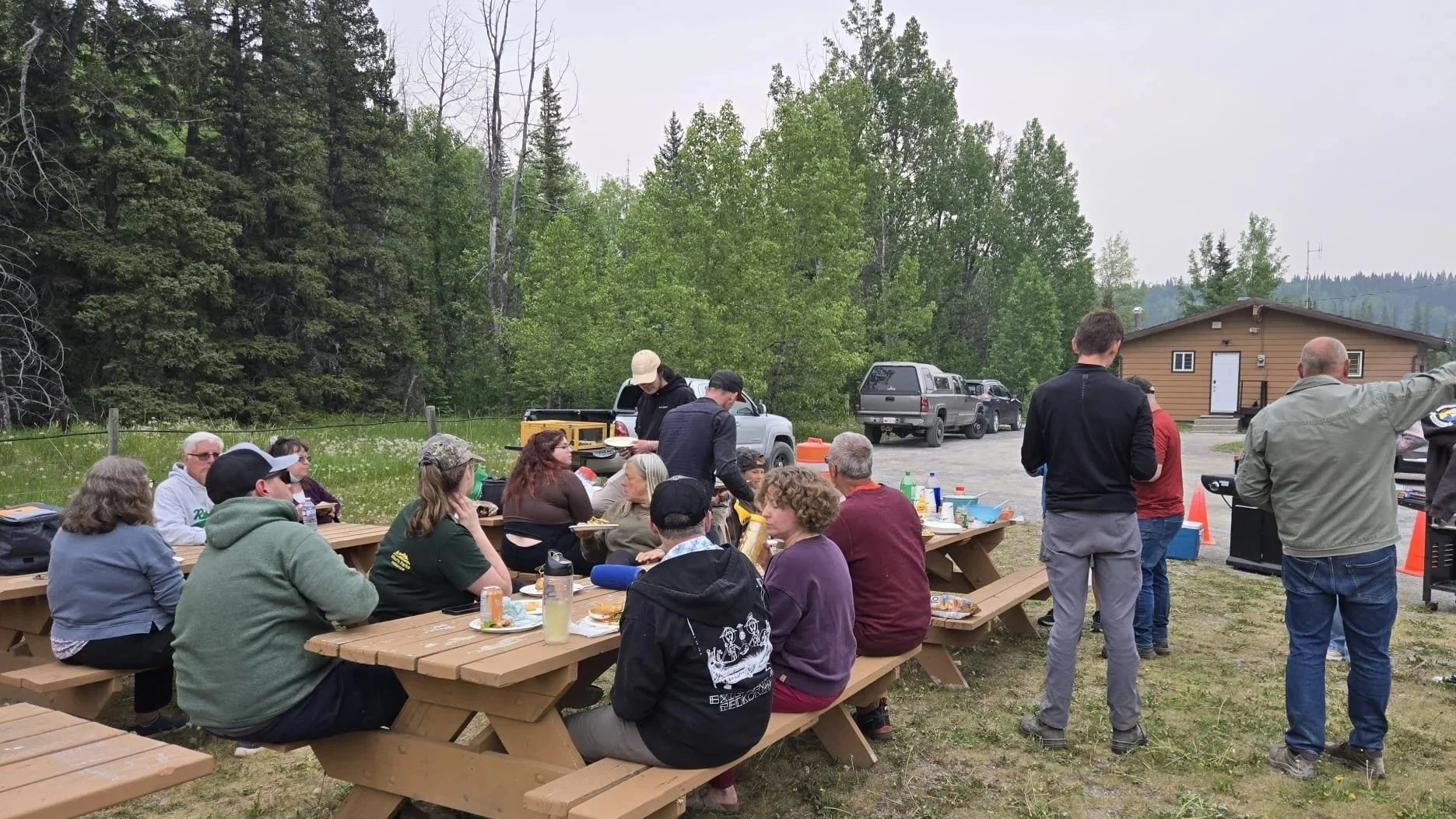 A group of people gathered outdoors for a meal at picnic tables, with cars and a small building in the background, surrounded by trees.
