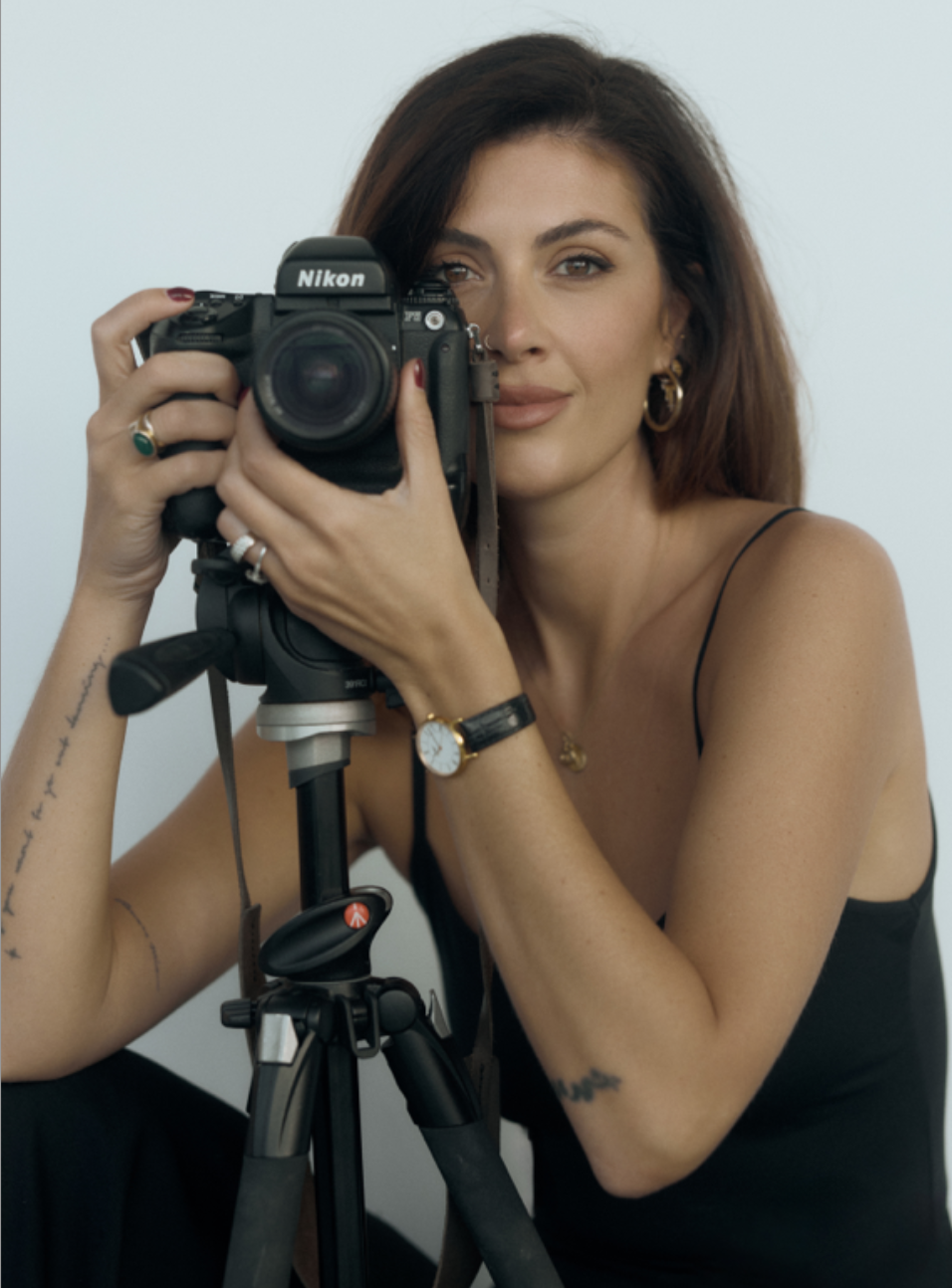 A woman with brown hair and wearing a black dress, holding a Nikon camera on a tripod, looking through the viewfinder.