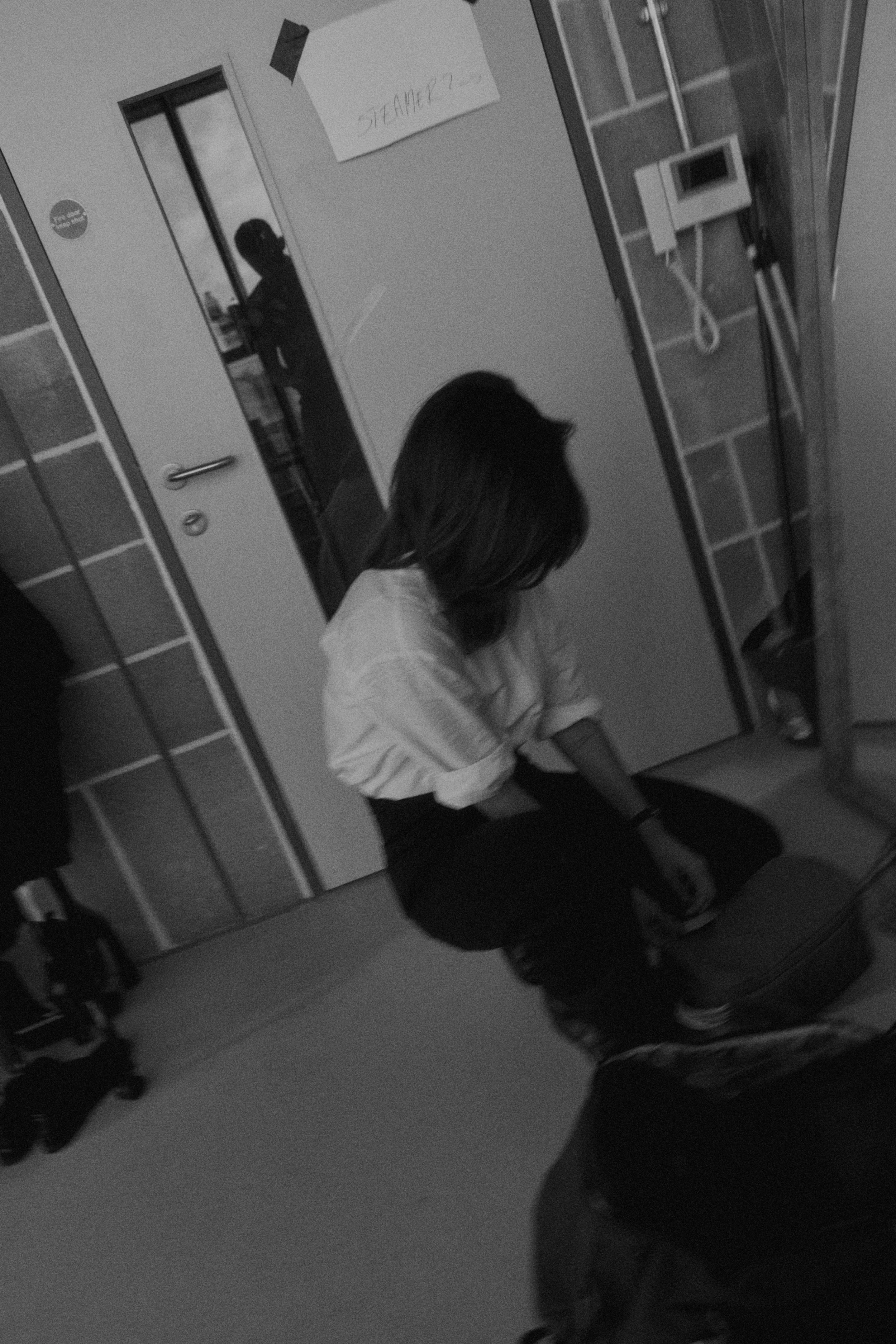A woman with shoulder-length hair wearing a white shirt and black pants, sitting on the floor in front of a door with a mirror and a suitcase nearby.