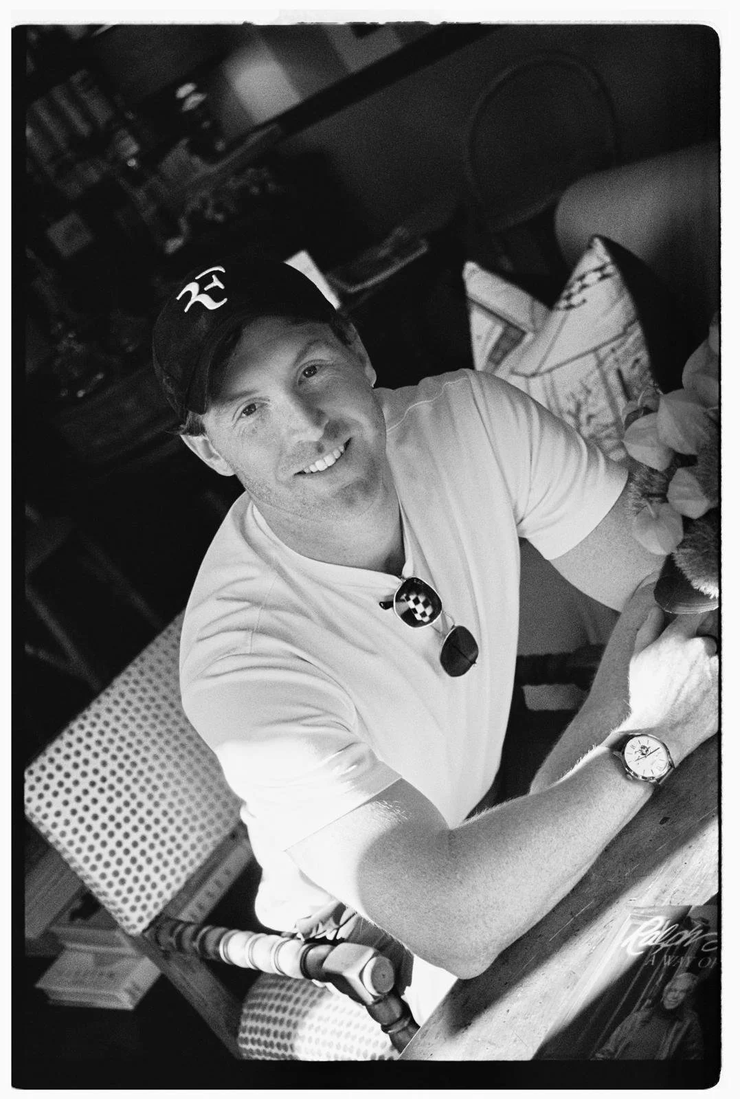 A smiling man wearing a black cap, white T-shirt, and a wristwatch, sitting at a table in a cozy indoor space with bookshelves and various items in the background.