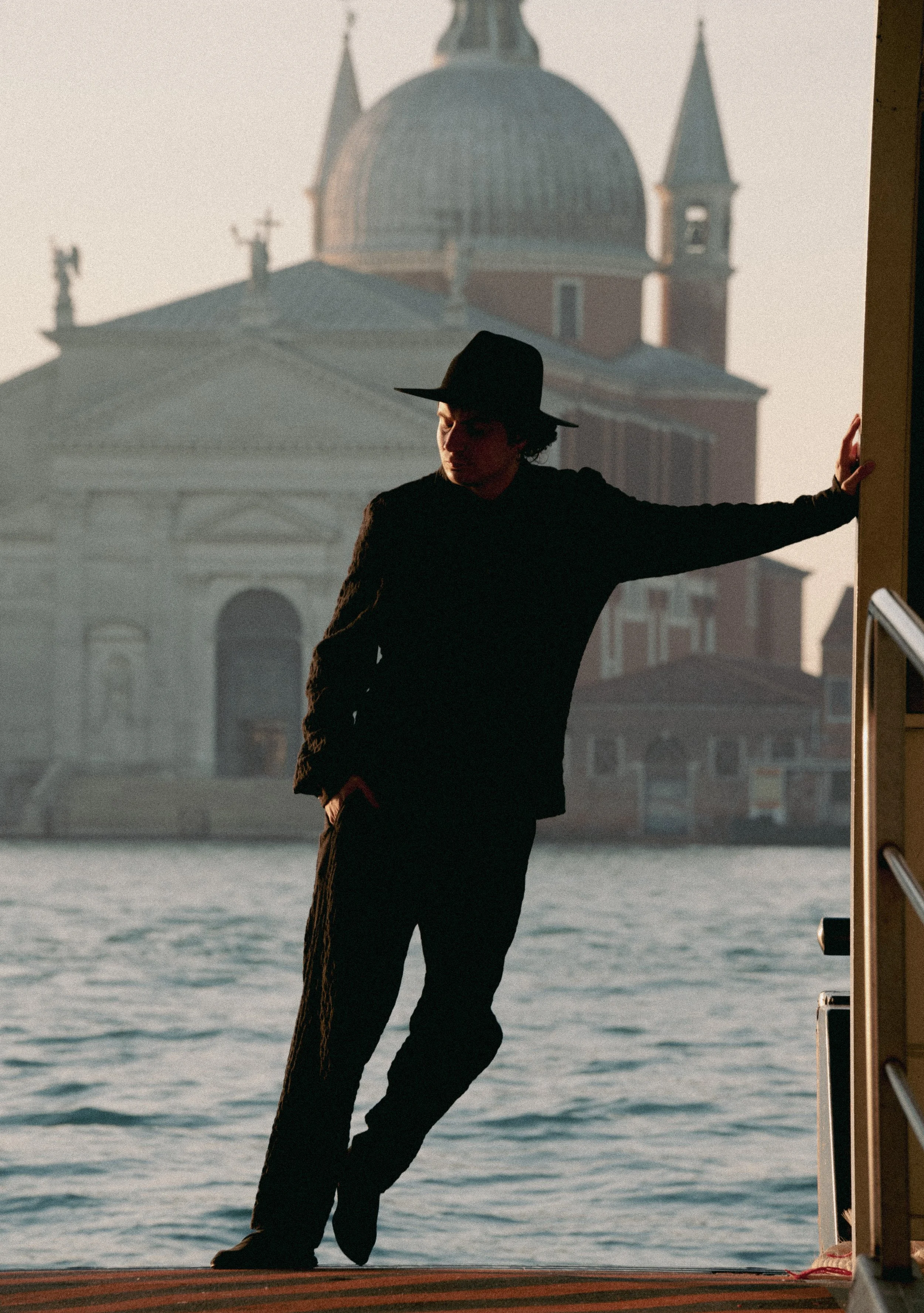 A person in dark clothing and a wide-brimmed hat stands by a waterfront with a historic building in the background, possibly in Venice, Italy.