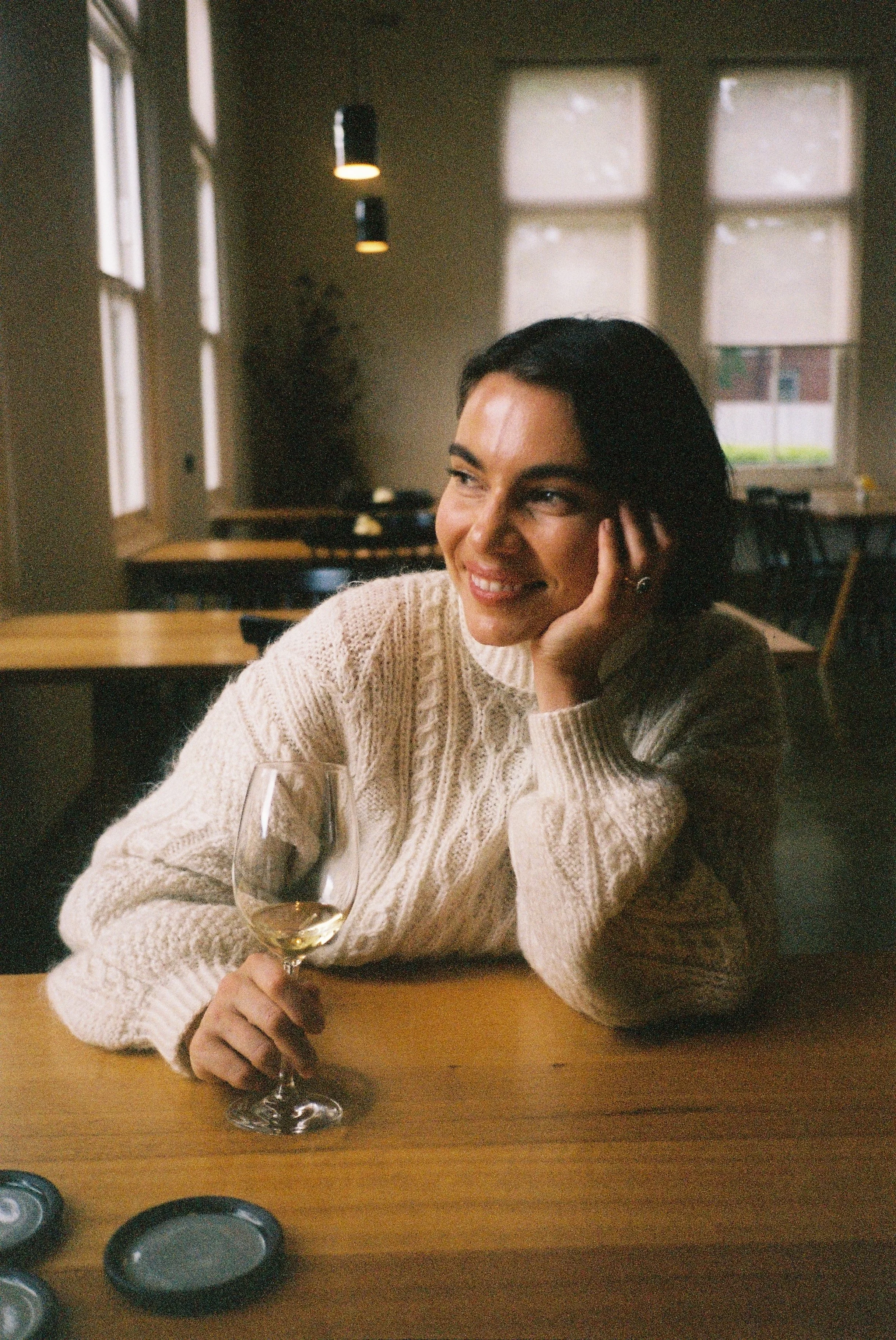 A woman with short dark hair, wearing a cream cable-knit sweater, sitting at a wooden table in a cozy, well-lit room with large windows and pendant lights. She is smiling, holding a glass of white wine, and resting her head on her hand.