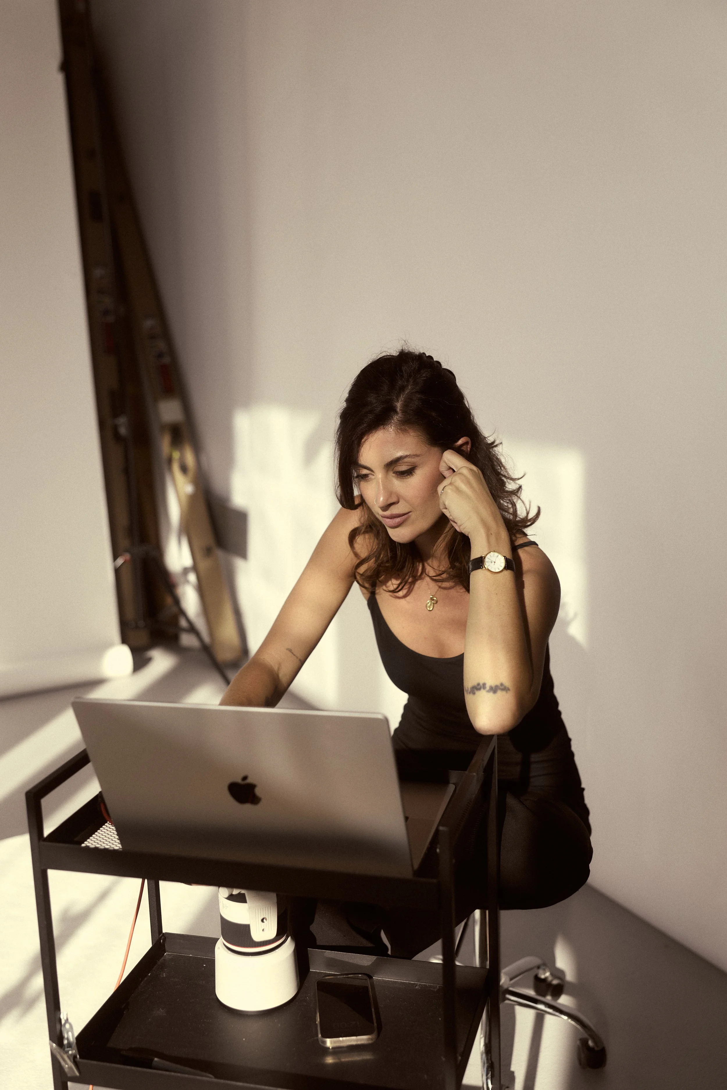 A woman with dark hair, wearing a black tank top, sitting at a small black table with a MacBook, coffee cup, and phone in a studio with plain white backdrop.