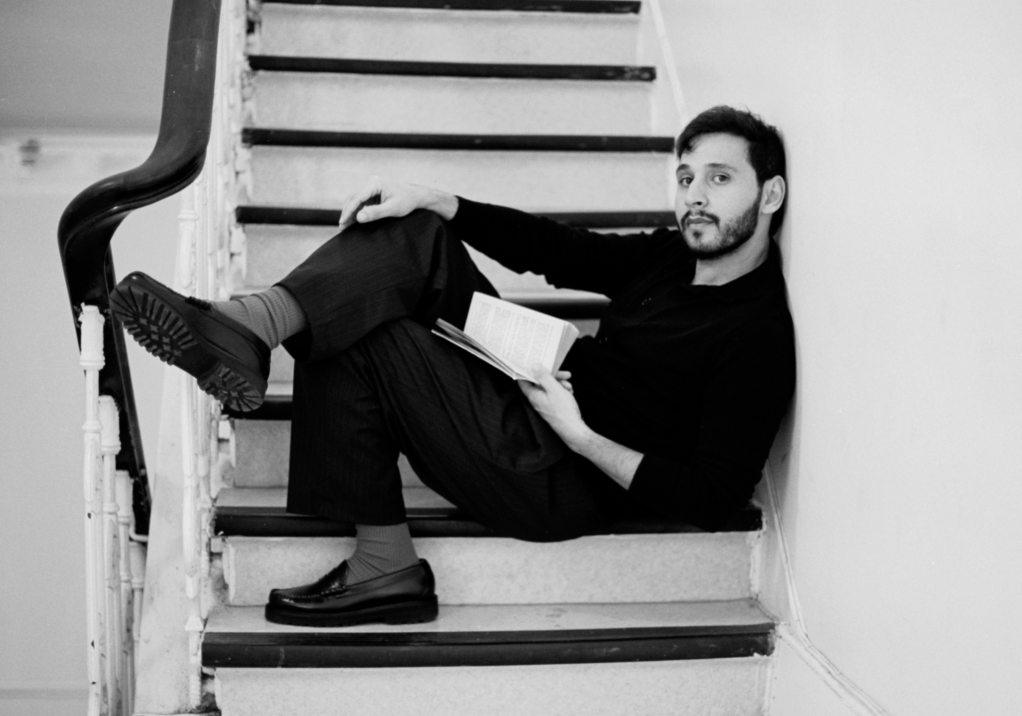 A man with dark hair, beard, wearing a black shirt, and striped pants, sitting on a staircase, reading a book, with one leg crossed over the other, and looking at the camera.