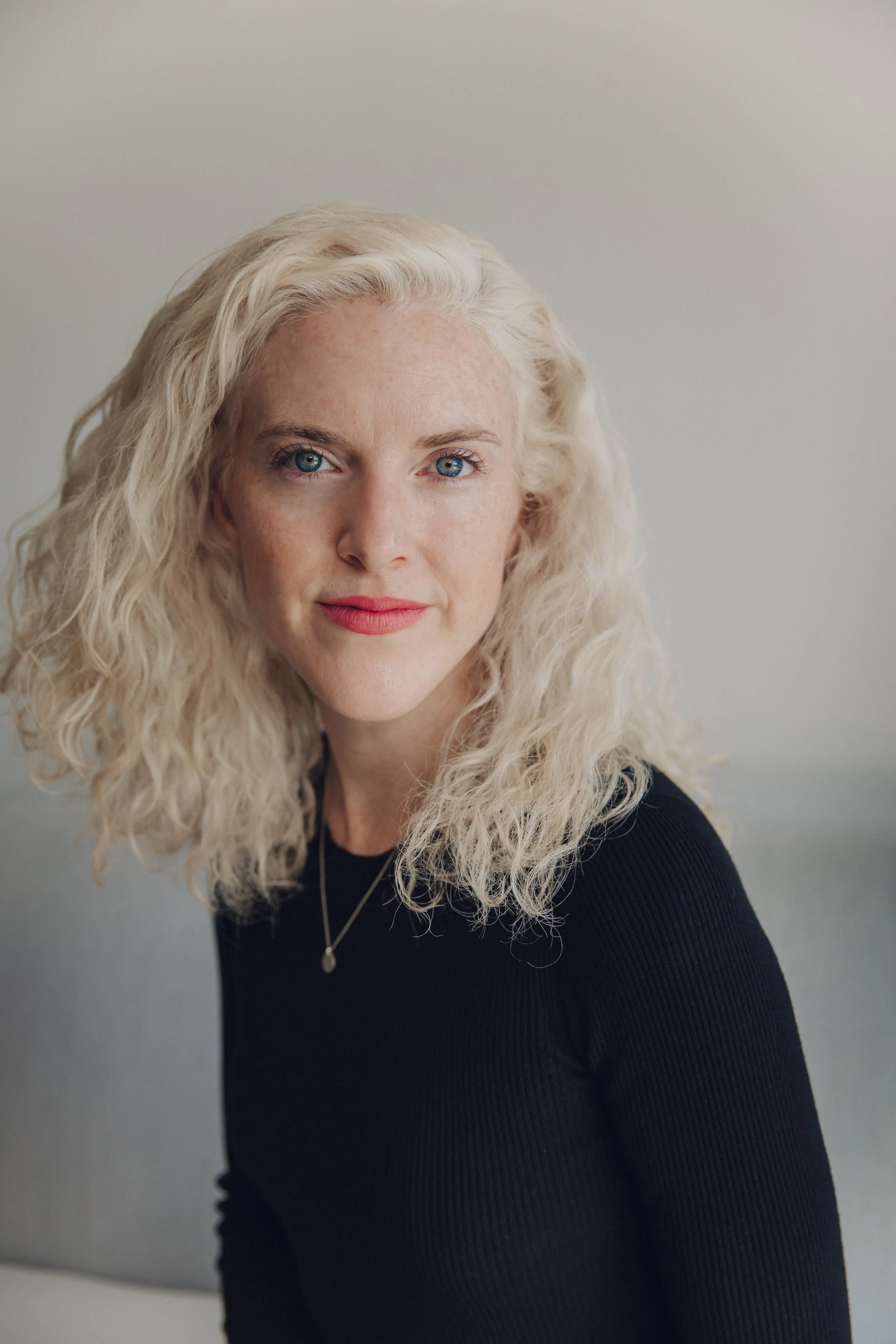 A woman with long, curly blonde hair, blue eyes, and wearing a black top, posing against a plain light-colored background.