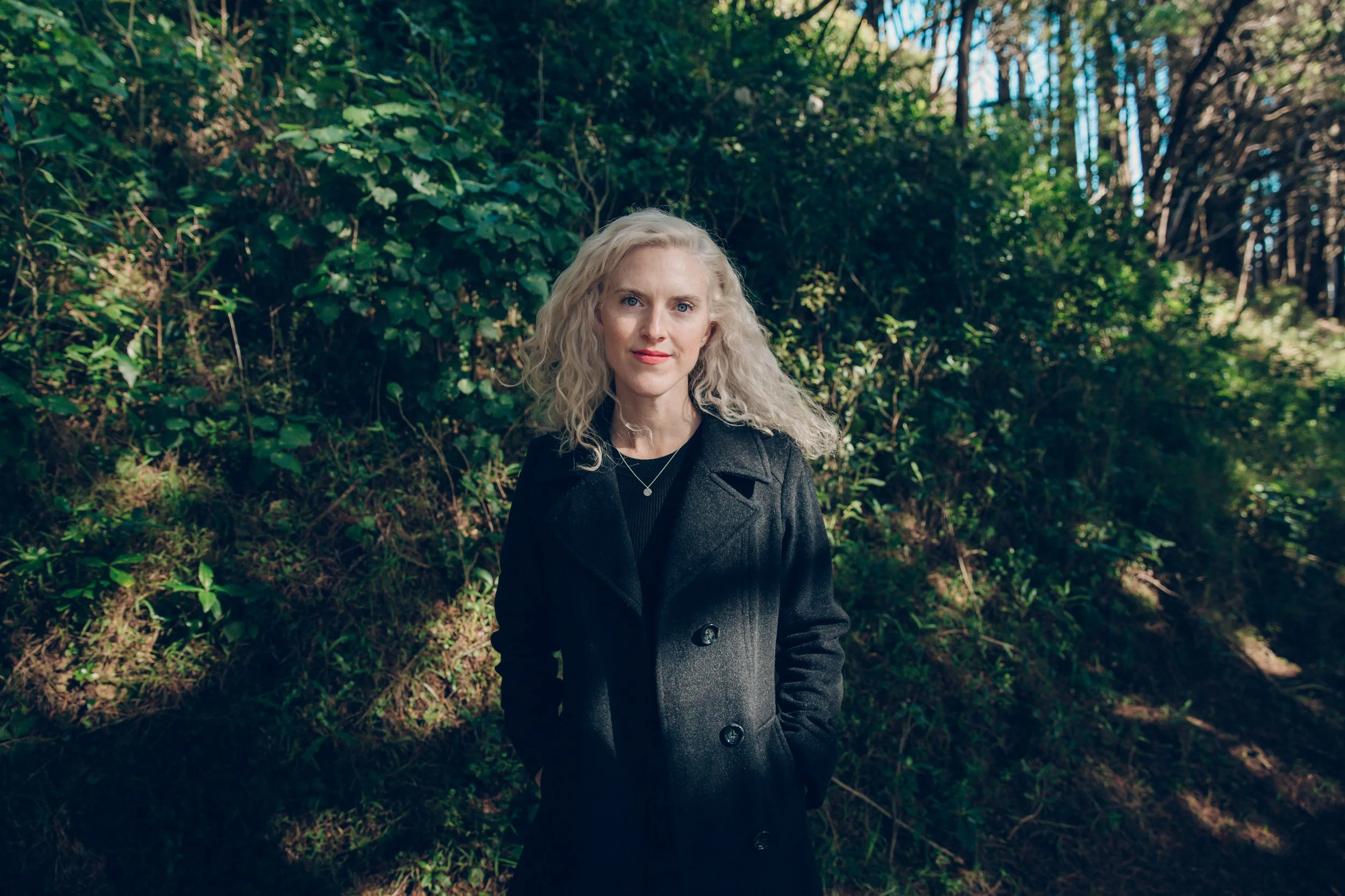 A woman with light curly hair standing outdoors in front of green bushes and trees, wearing a black coat and a small necklace.